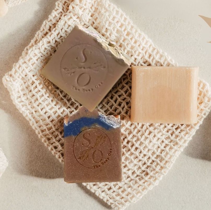 Three bars of soap in different colors rest on a beige mesh bag.