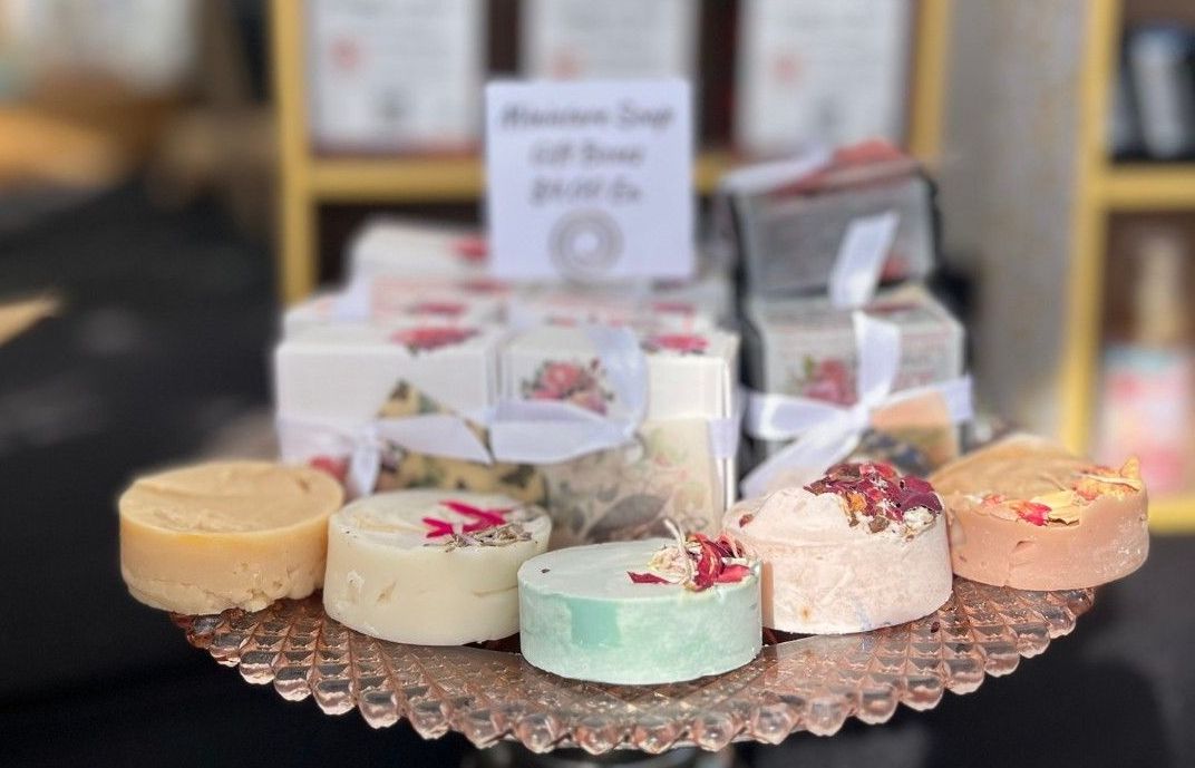 A display of handmade soaps on a glass cake stand. The soaps are round and square, in various colors, with floral accents.