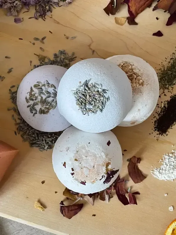 Four white bath bombs topped with dried herbs, set on a wooden surface. Surrounding ingredients like rose petals and lavender.