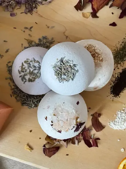 Four white bath bombs topped with dried herbs, set on a wooden surface. Surrounding ingredients like rose petals and lavender.
