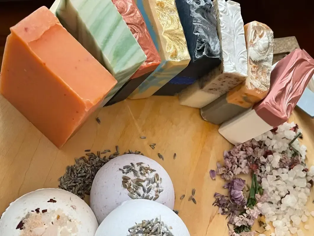 Assortment of colorful handmade soaps and bath bombs arranged on a wooden surface, sprinkled with dried lavender and salt.