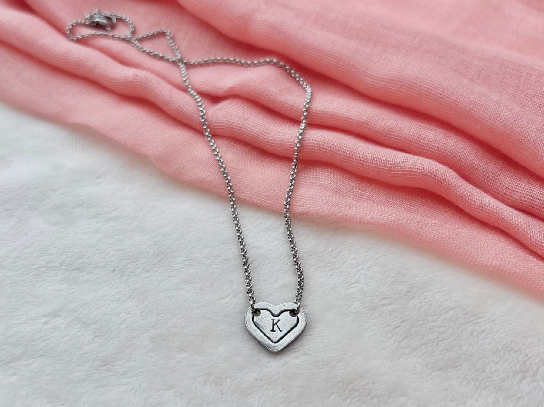 Silver necklace with a heart-shaped pendant, set against a pink cloth background.