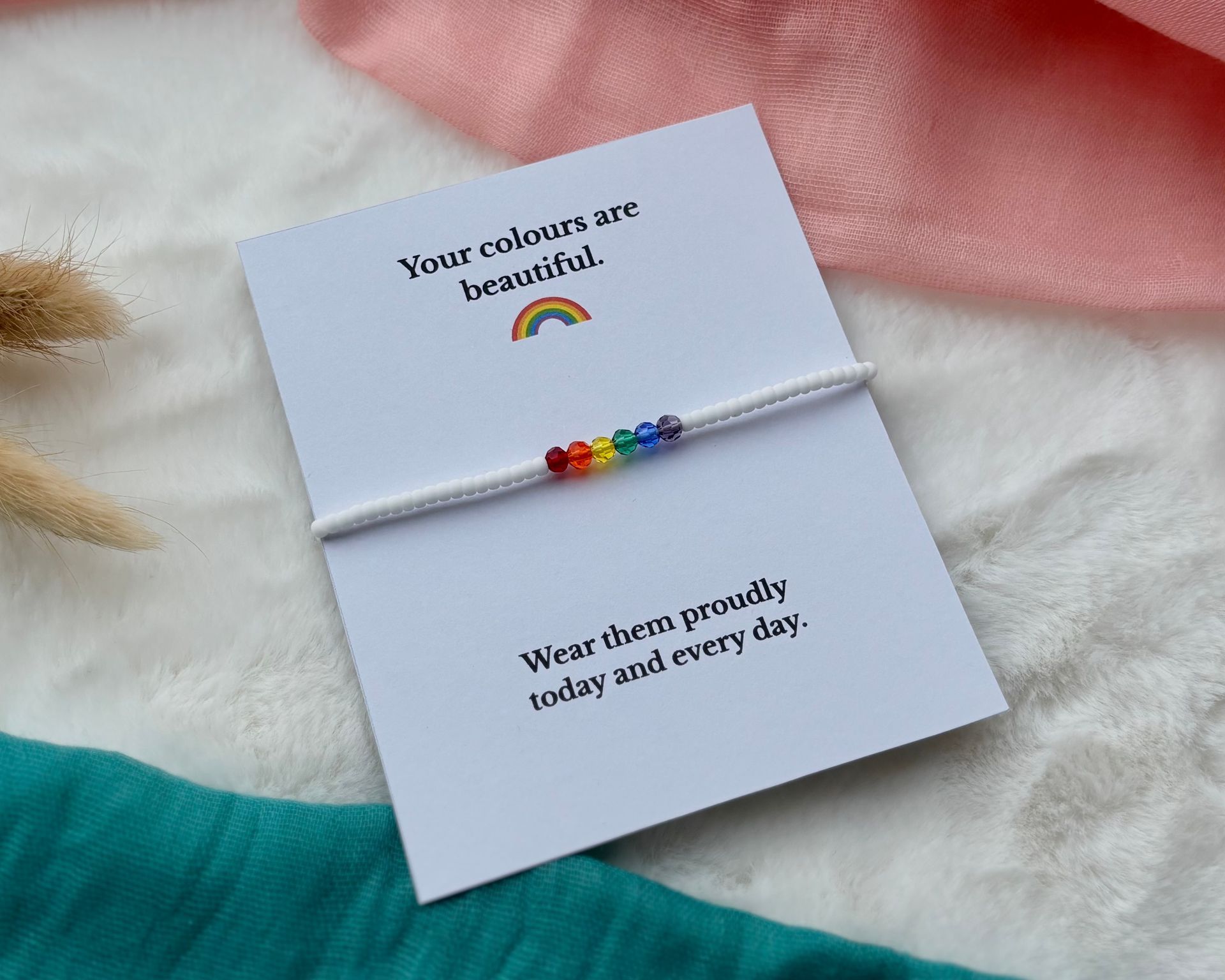 A bracelet with small rainbow-colored beads on a white card that reads, "Your colours are beautiful. Wear them proudly."