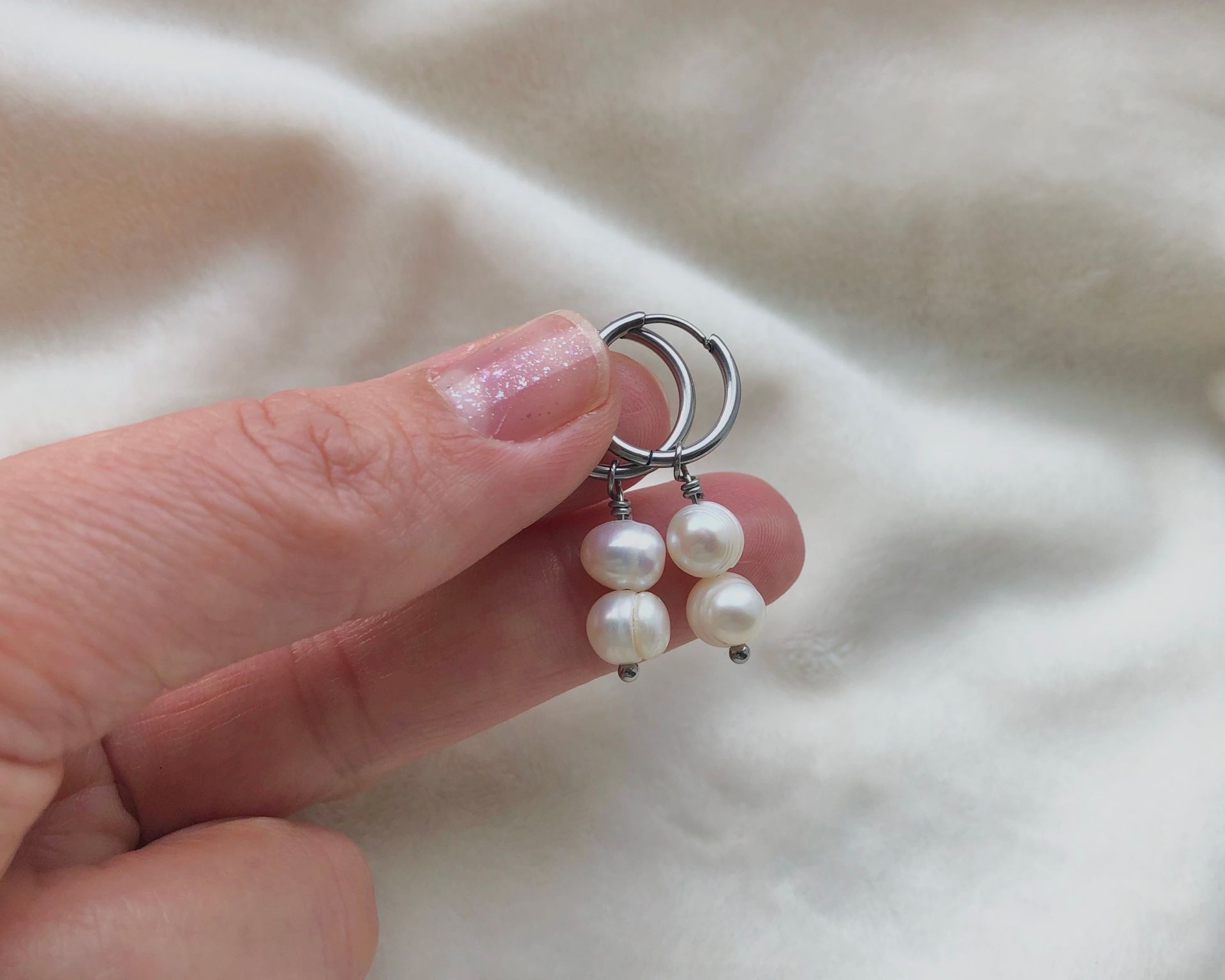 Hand holding a pair of white pearl earrings with silver hoops against a white cloth.