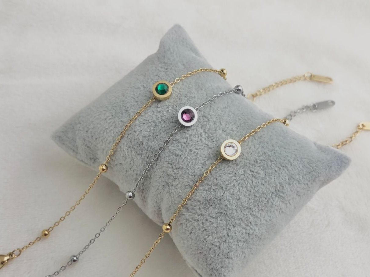Three delicate bracelets with colorful gems on a gray cushion.