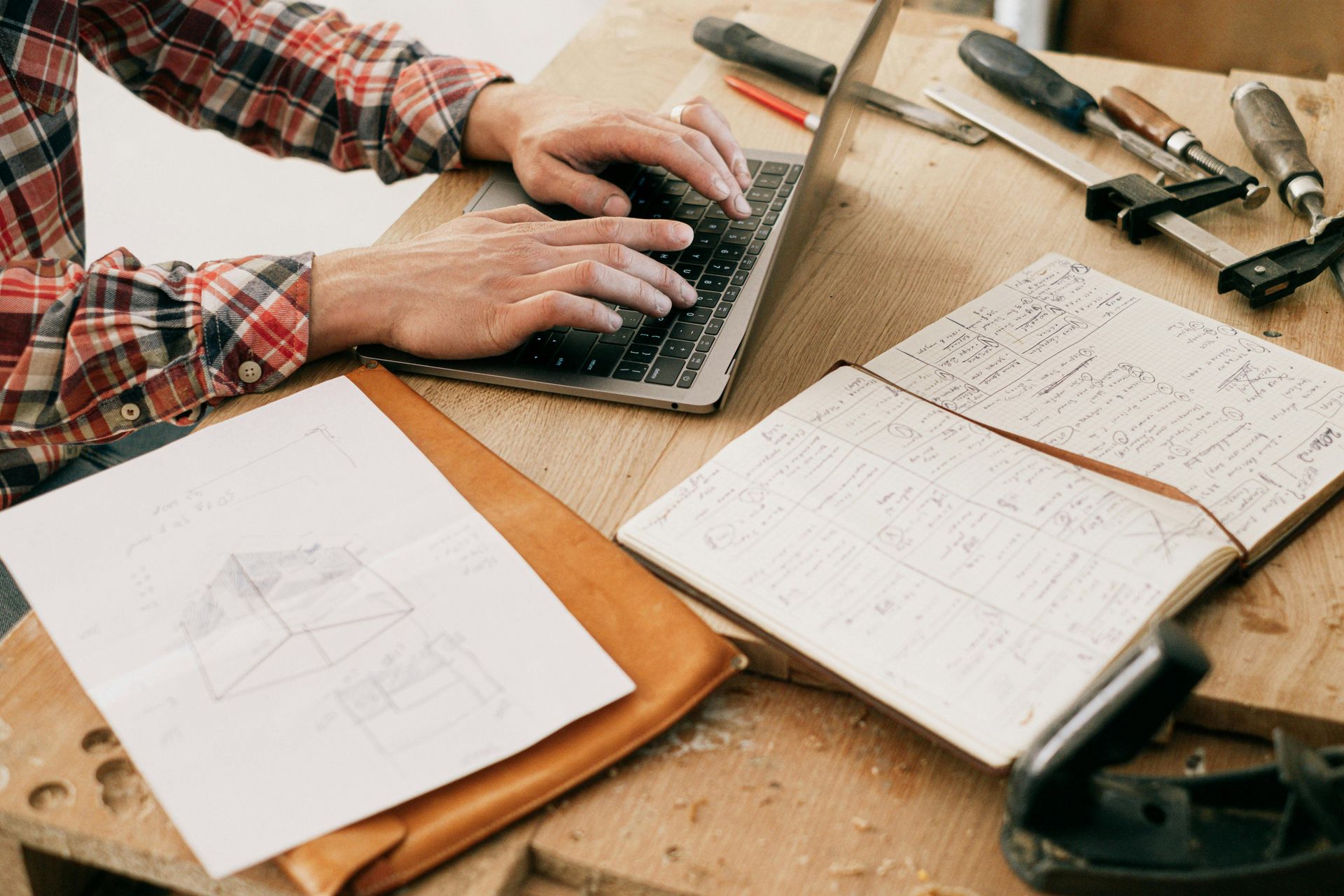 Hands typing on laptop at a woodworking table with sketches, tools, and notebook.