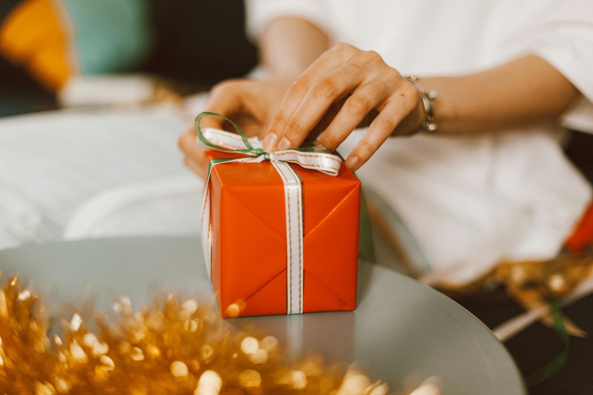 Hands tying a ribbon on a wrapped red gift box, set on a table with gold tinsel in the foreground.