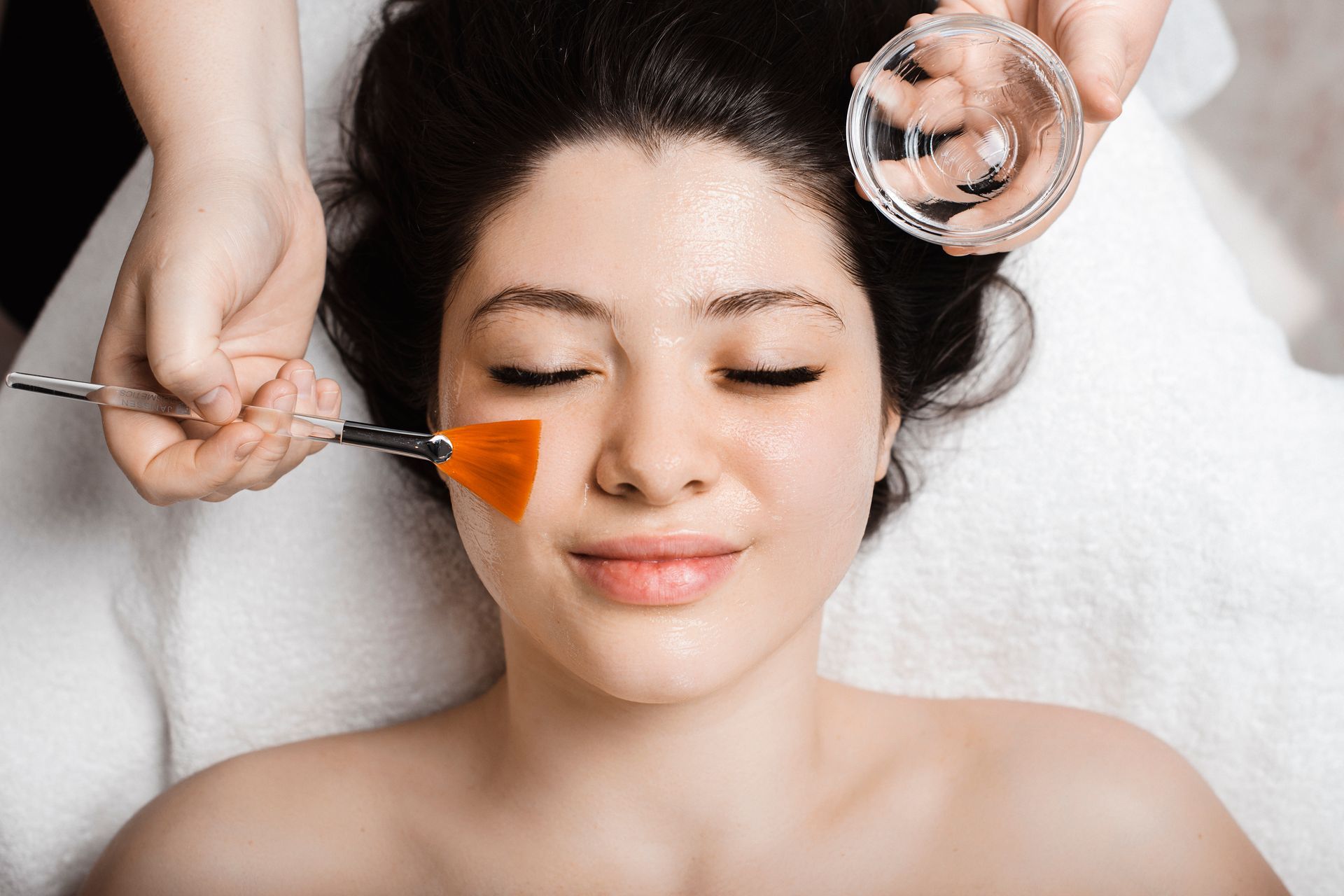Facial chemical peel application. A person lies with eyes closed as esthetician applies product with a brush.