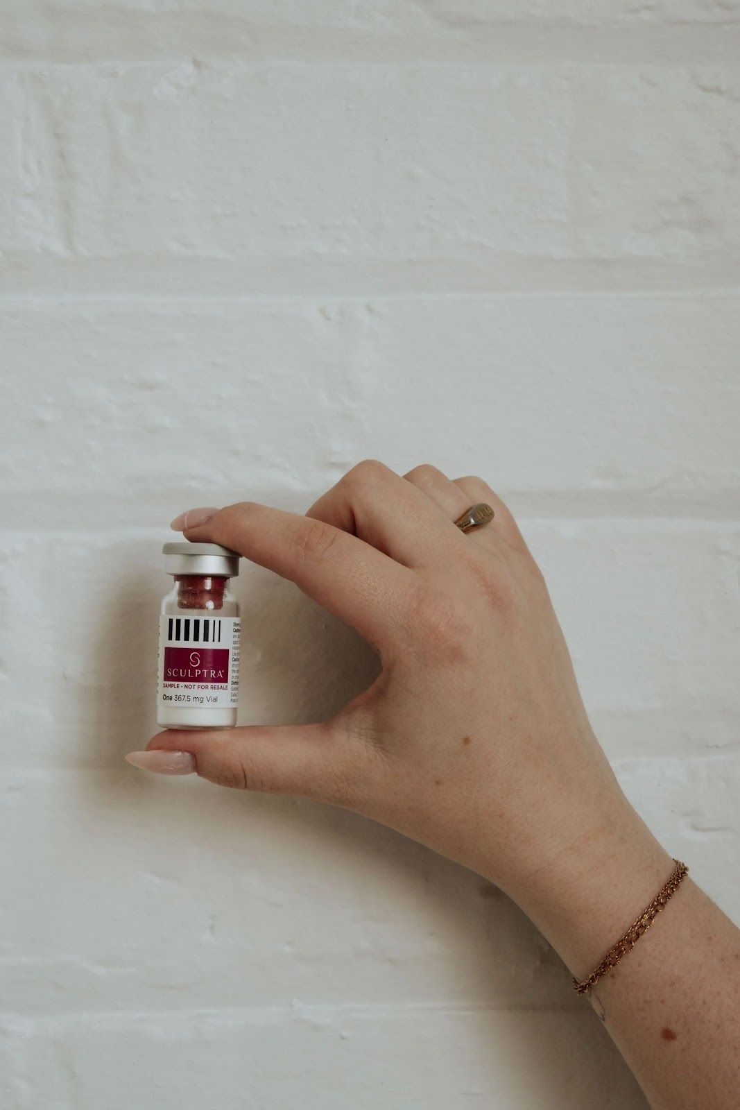 A hand holds a small glass vial with a white and magenta label against a white brick wall.
