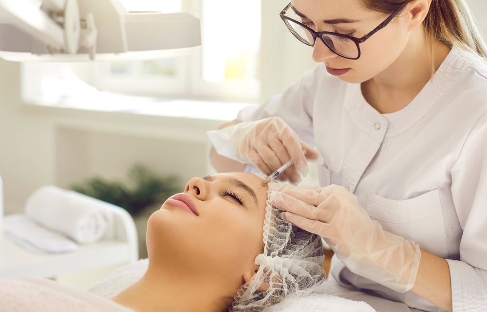 A practitioner in a white coat and gloves administering a facial injection to a client lying on a treatment table.