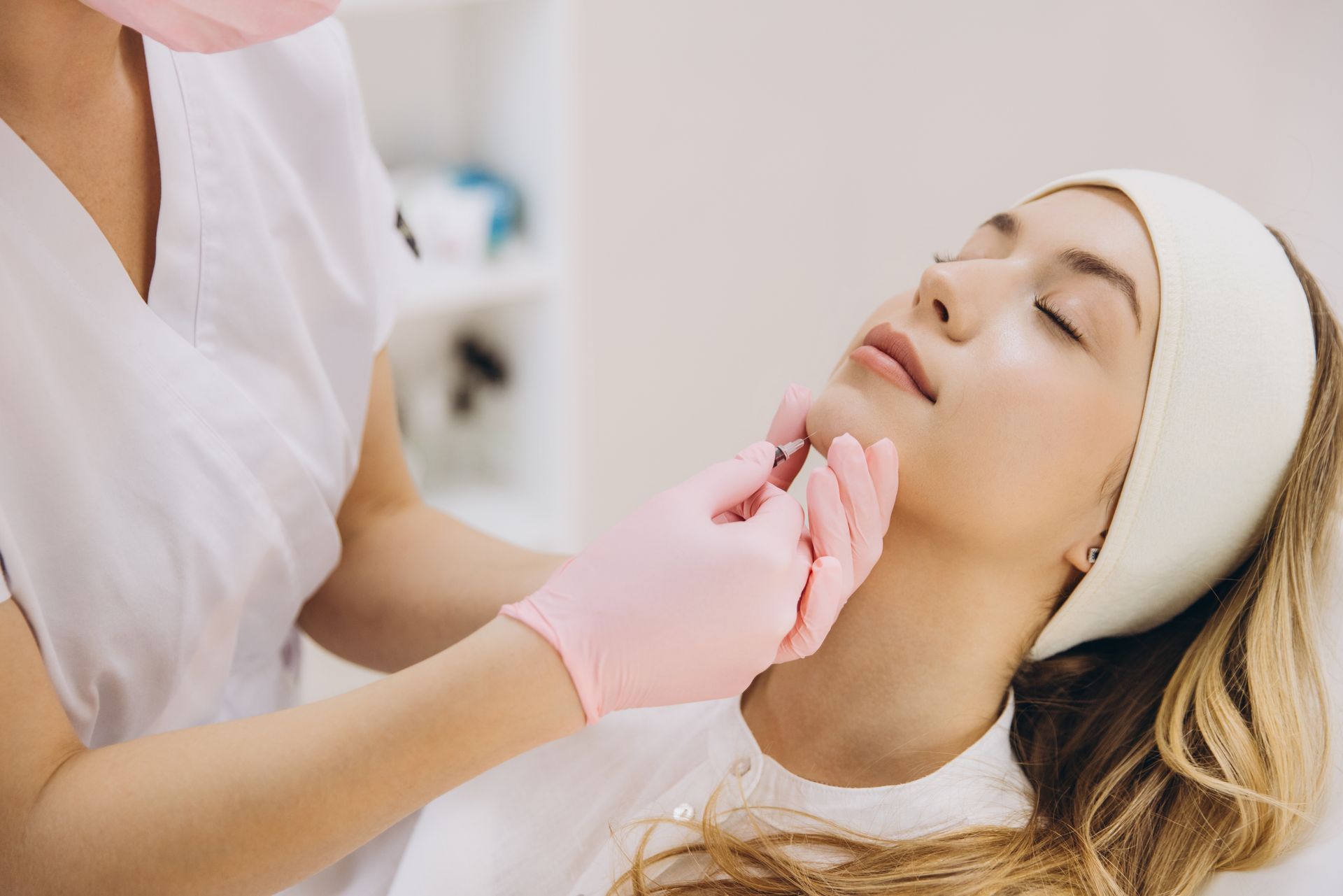 Woman receiving a facial injection, performed by a person wearing pink gloves and a mask, in a clinical setting.