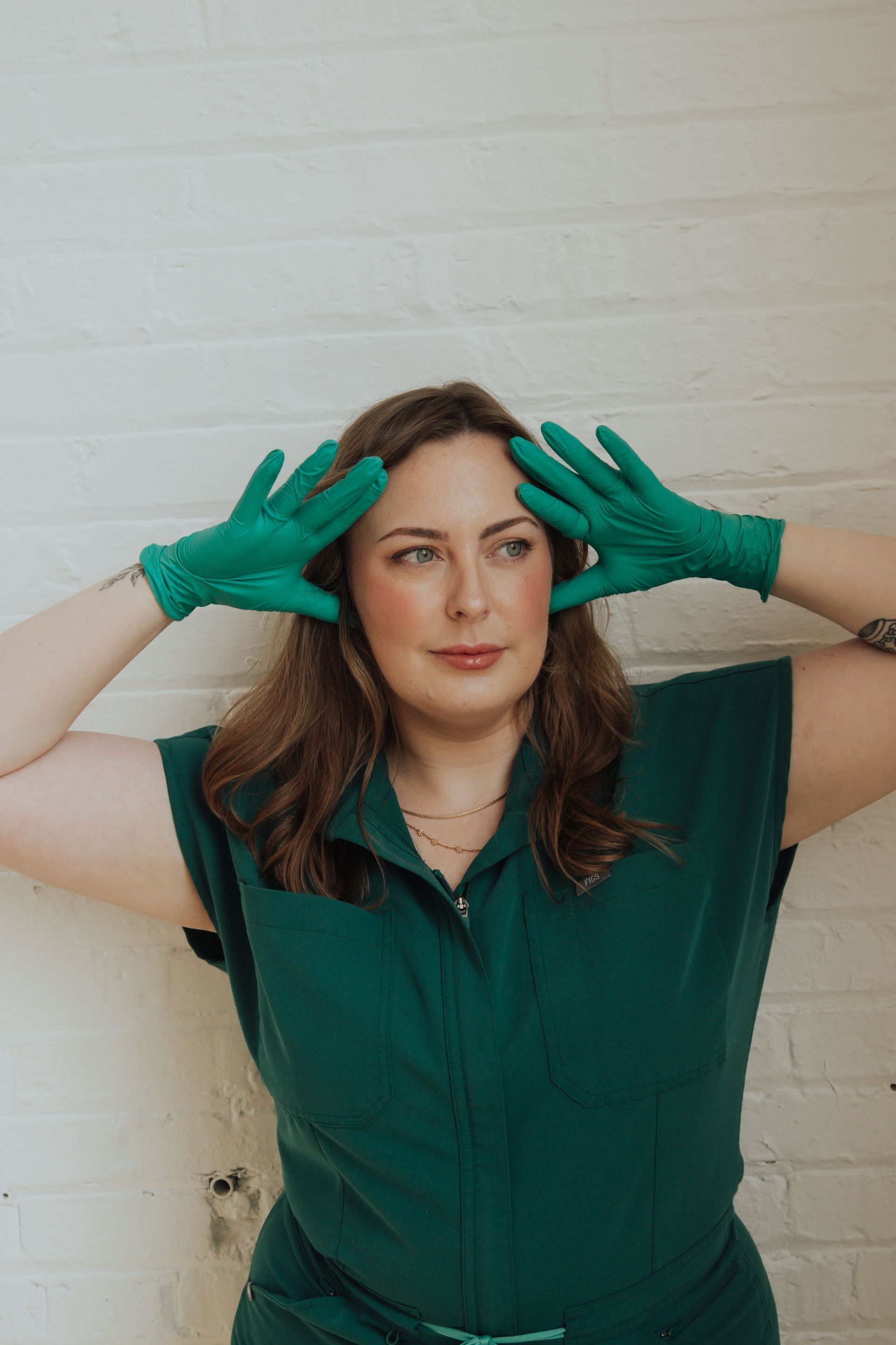 Woman in green jumpsuit and gloves, hands near head, leaning against a white brick wall.