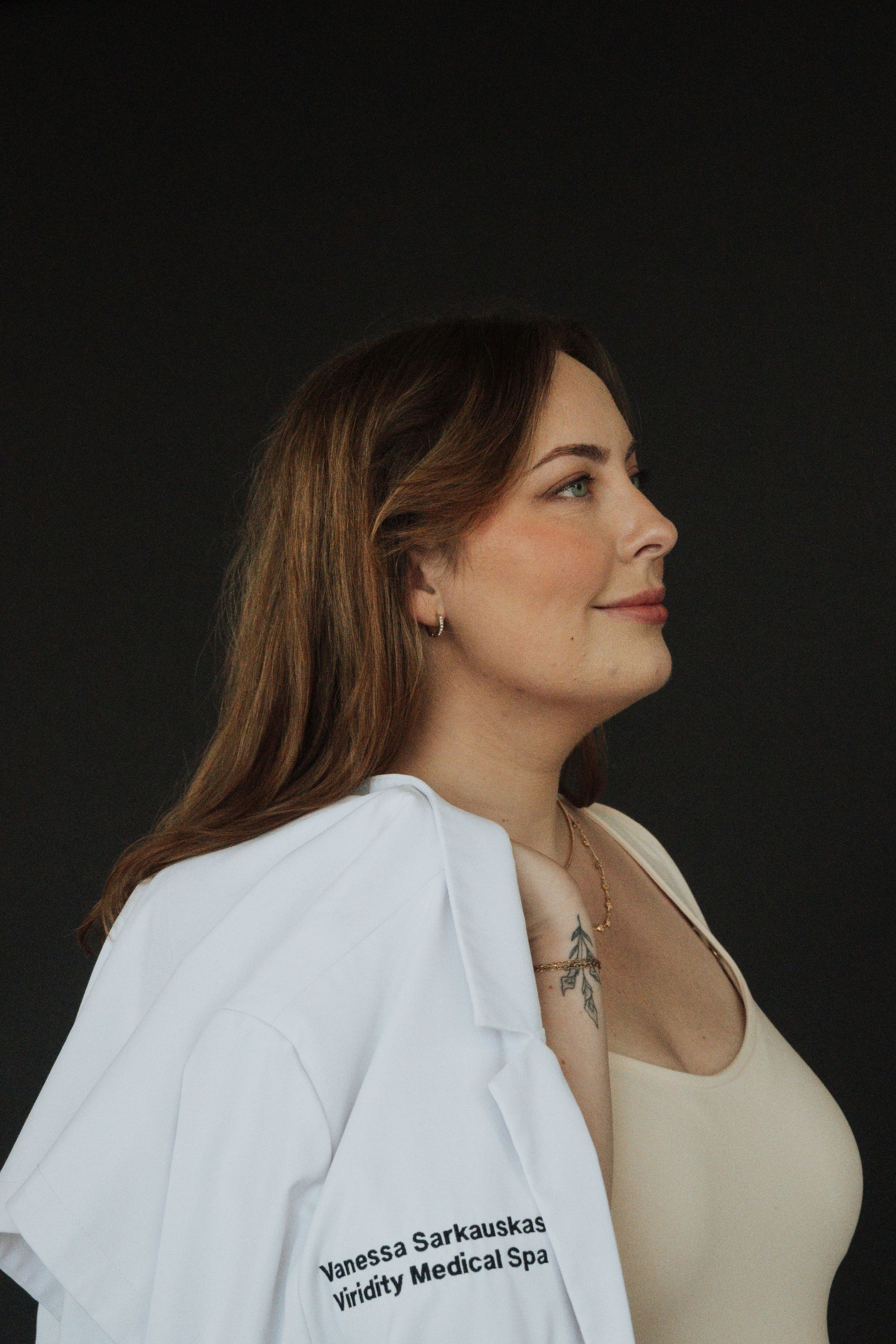 Woman with brown hair and a medical coat, smiling, looking to the right. Dark background.