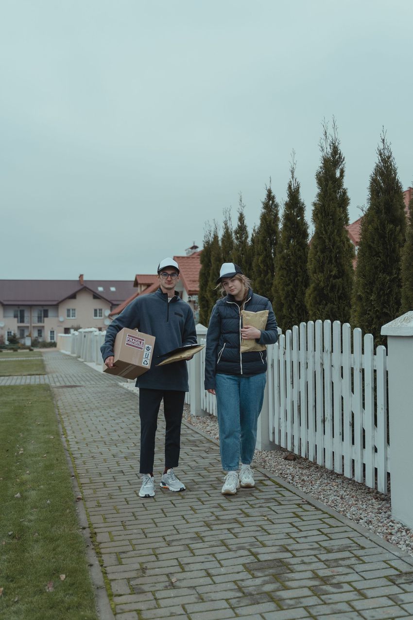 Two people walking on a cobblestone path beside a white fence, one holding a box and a cup.