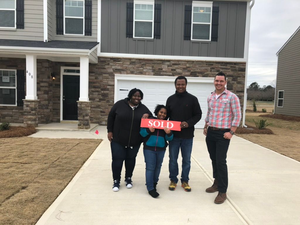Four people stand in front of a house holding a red โSOLDโ sign on the driveway.