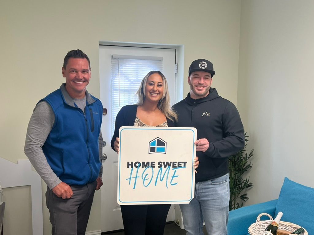 Three people in a room holding a โHome Sweet Homeโ sign, smiling beside a couch.