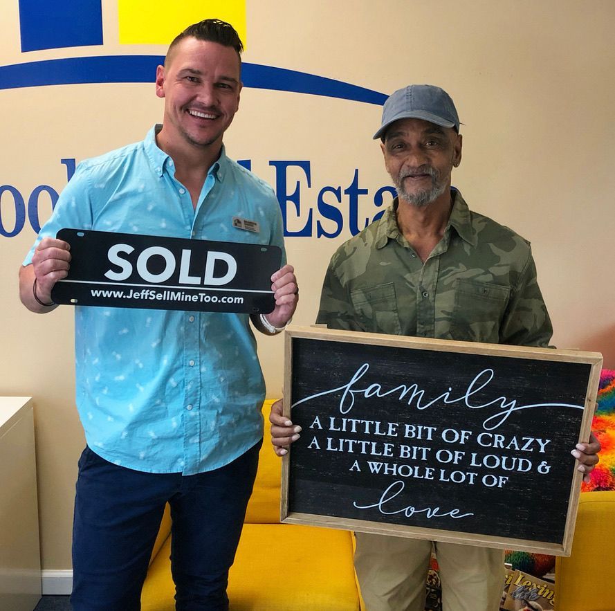 Two men hold real estate signs: โSOLDโ and โfamily...a little bit of crazy...โ in an office.