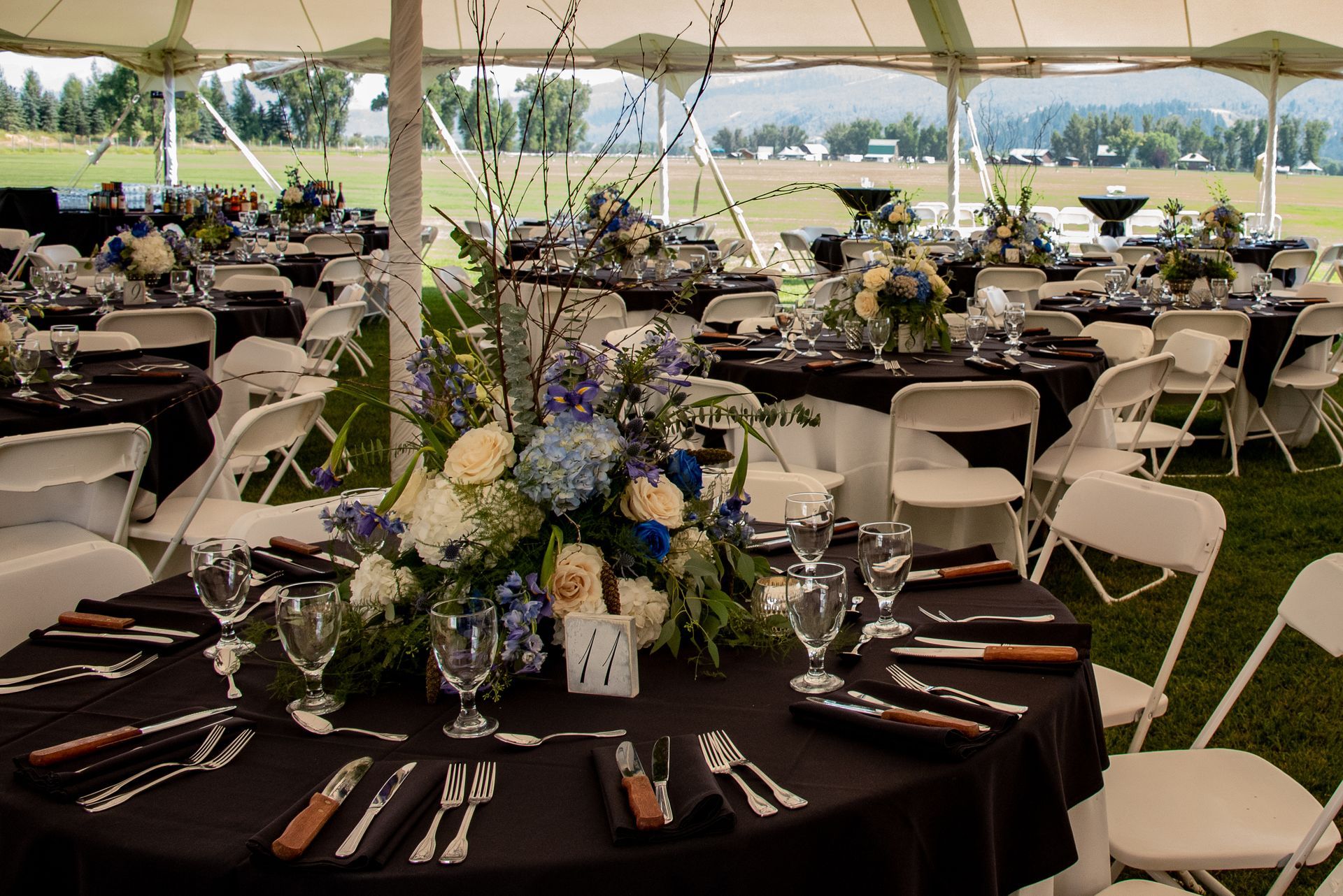 Outdoor tent set for a wedding reception with black-clothed round tables, white chairs, and floral centerpieces.