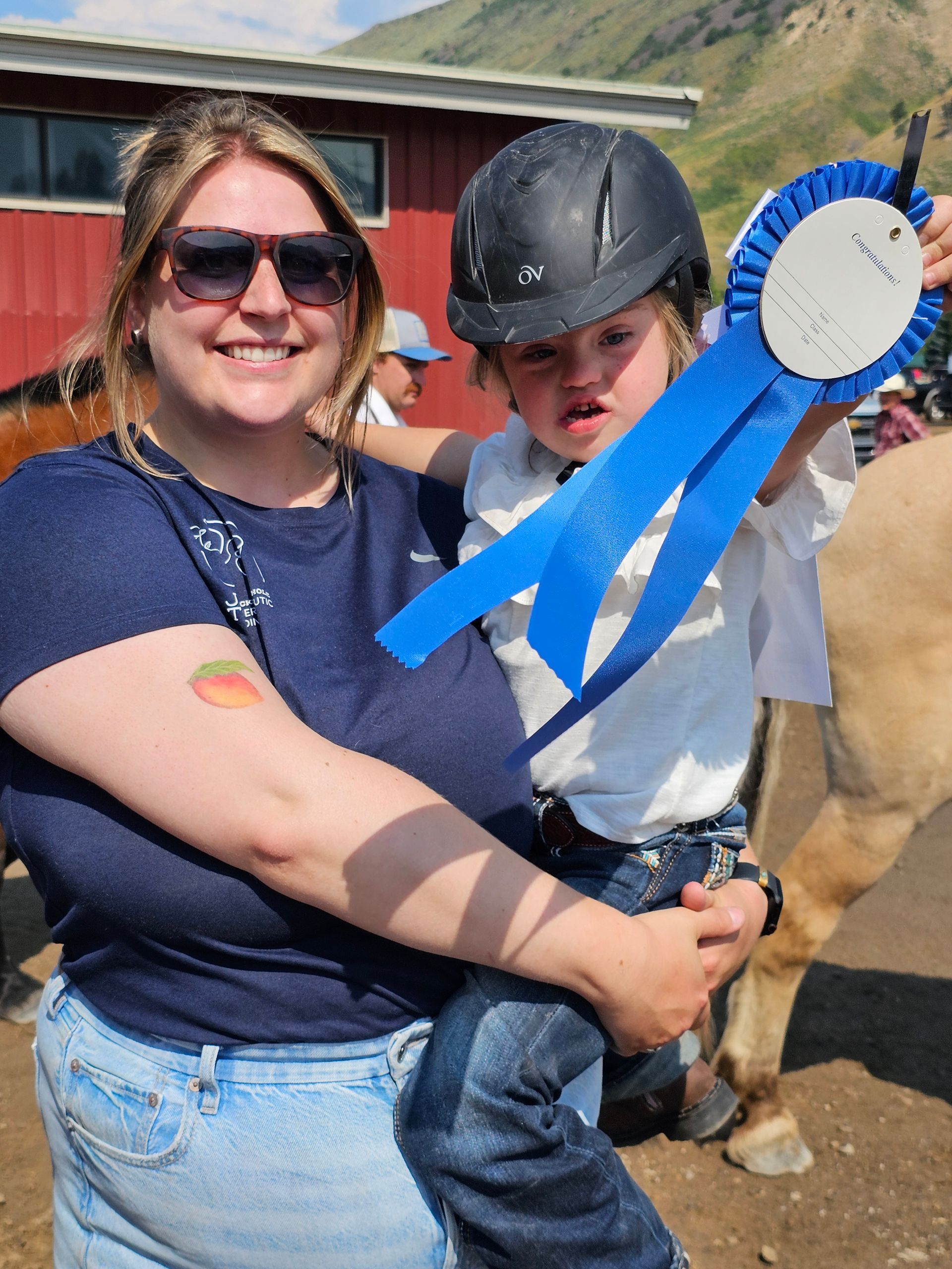 A person holding a child wearing a riding helmet and holding a blue award ribbon in front of a red building and horse.