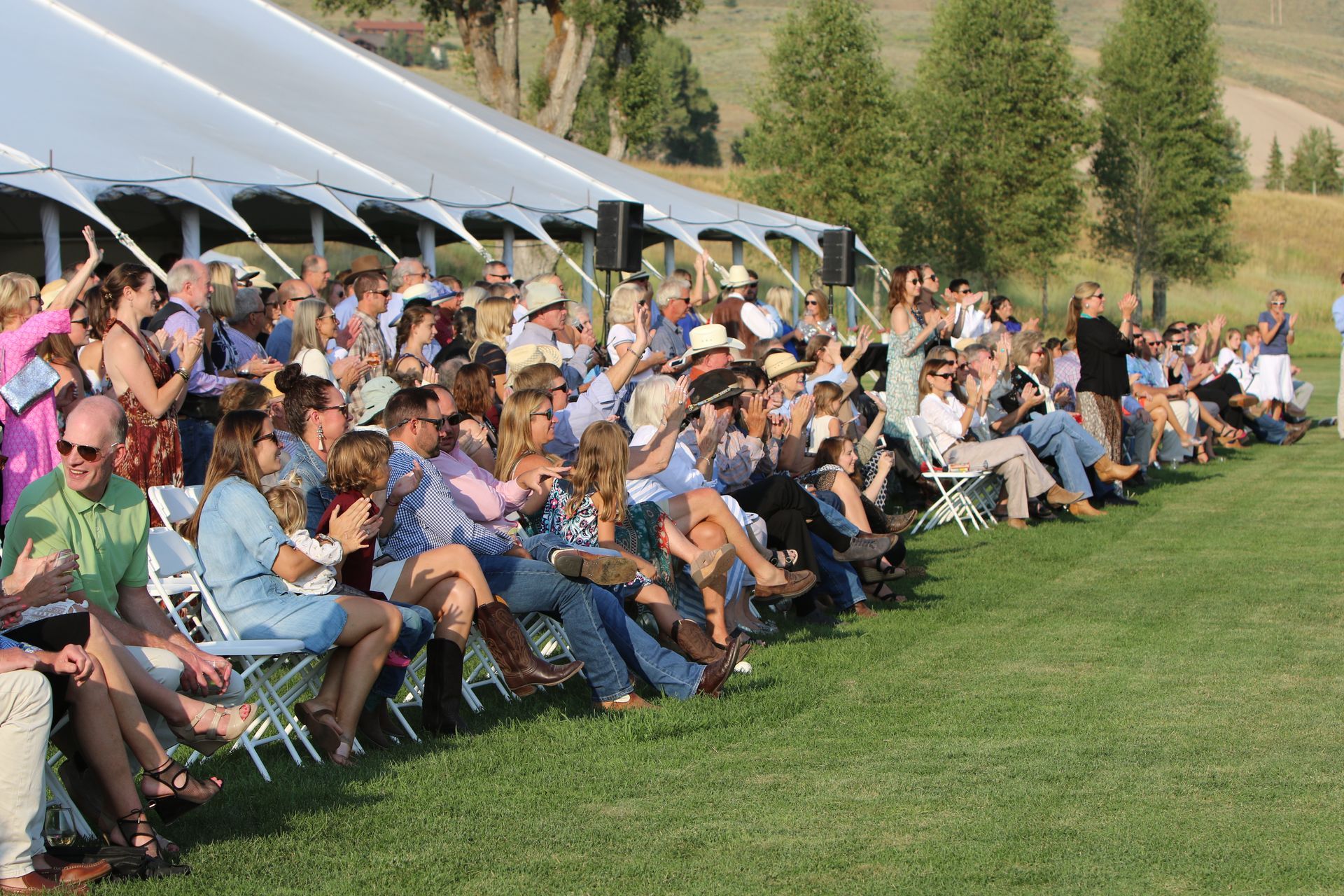 A large, diverse group of people sitting on chairs on a grassy field, watching a performance under a white event tent.