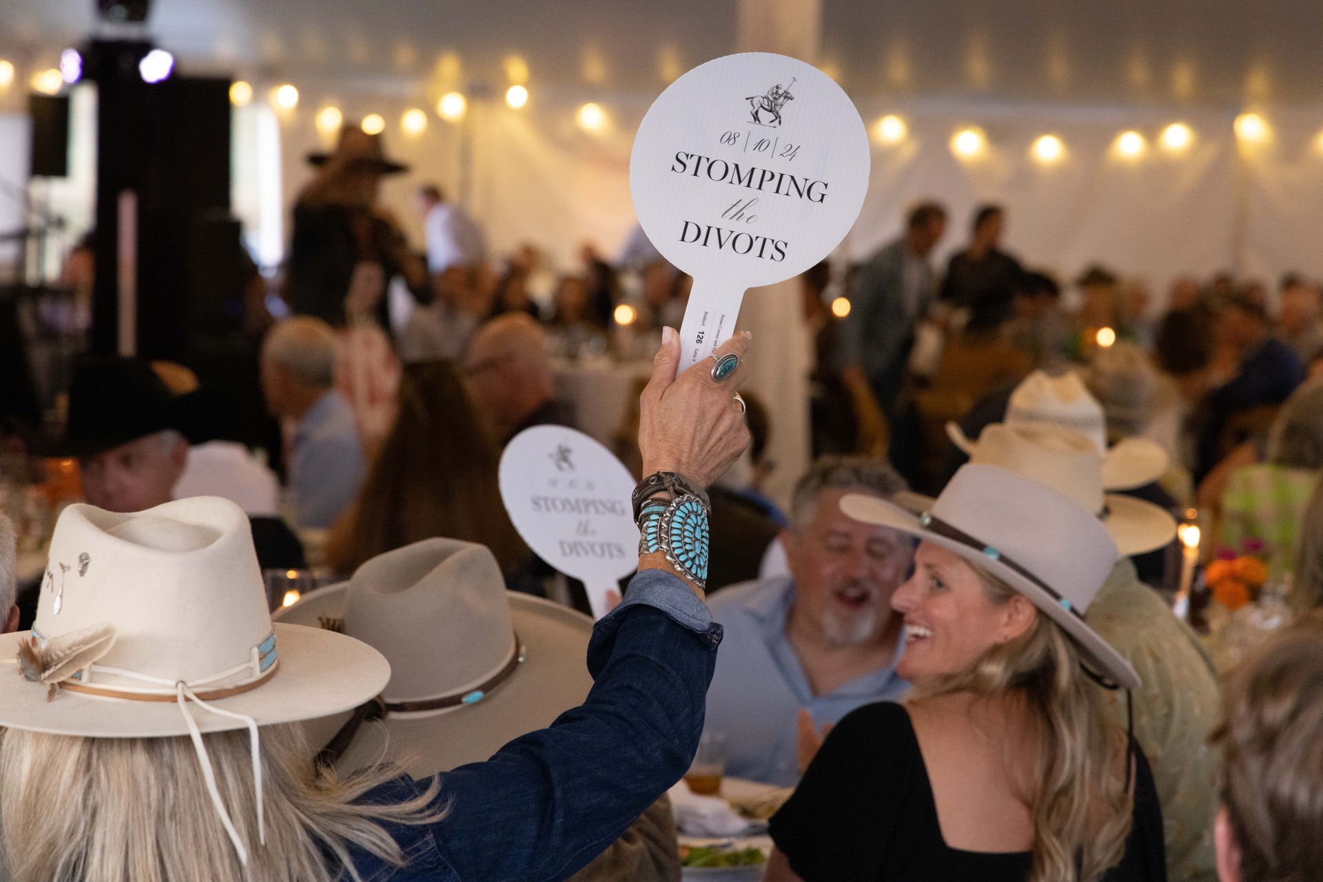 People in cowboy hats gather under a tent, one holding up a paddle reading 
