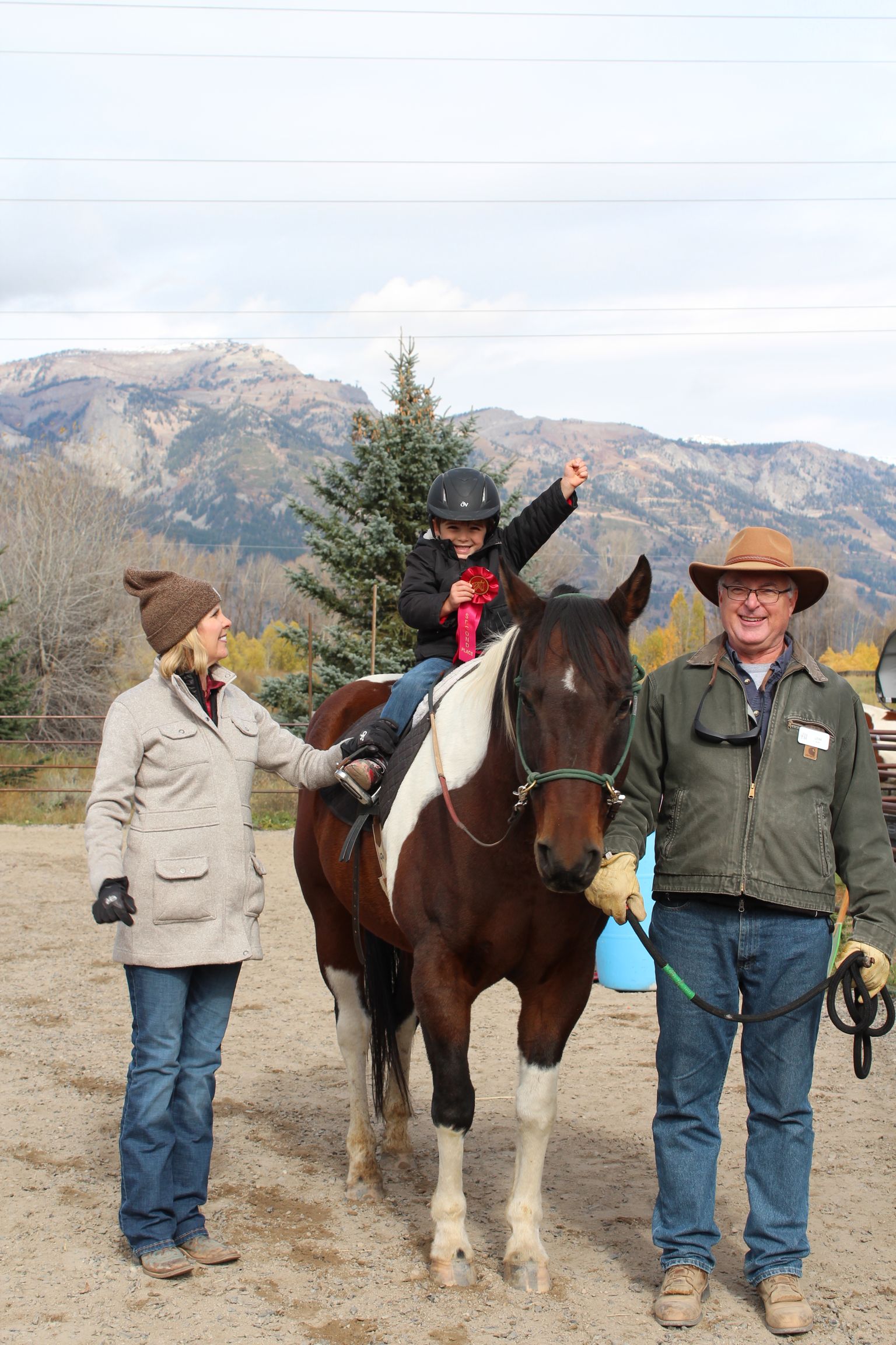 Man leading a black horse in a sandy arena. Man wears brown shirt and jeans.