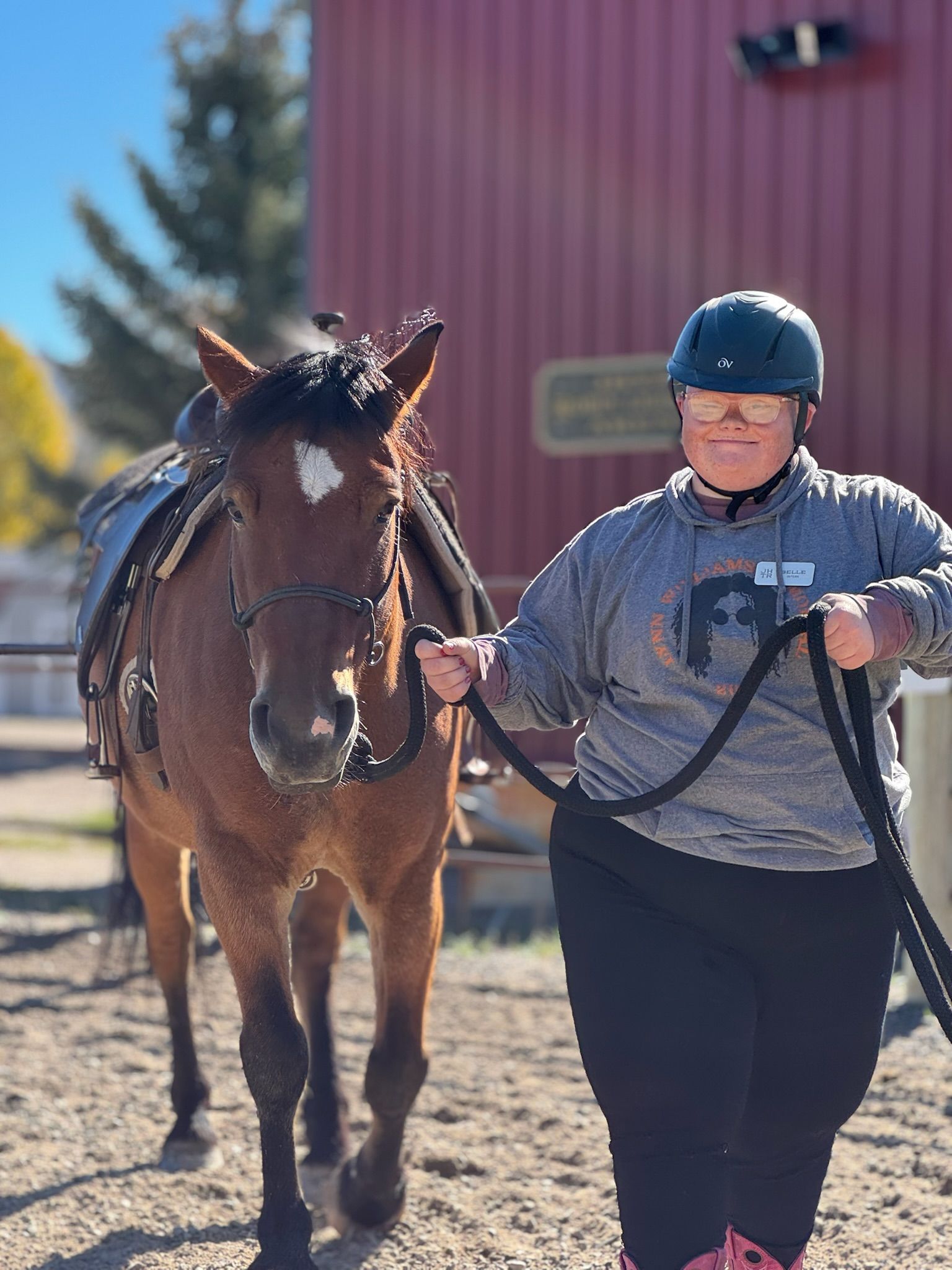 A child in helmet and a woman pet a brown horse outdoors by a red building; smiling.