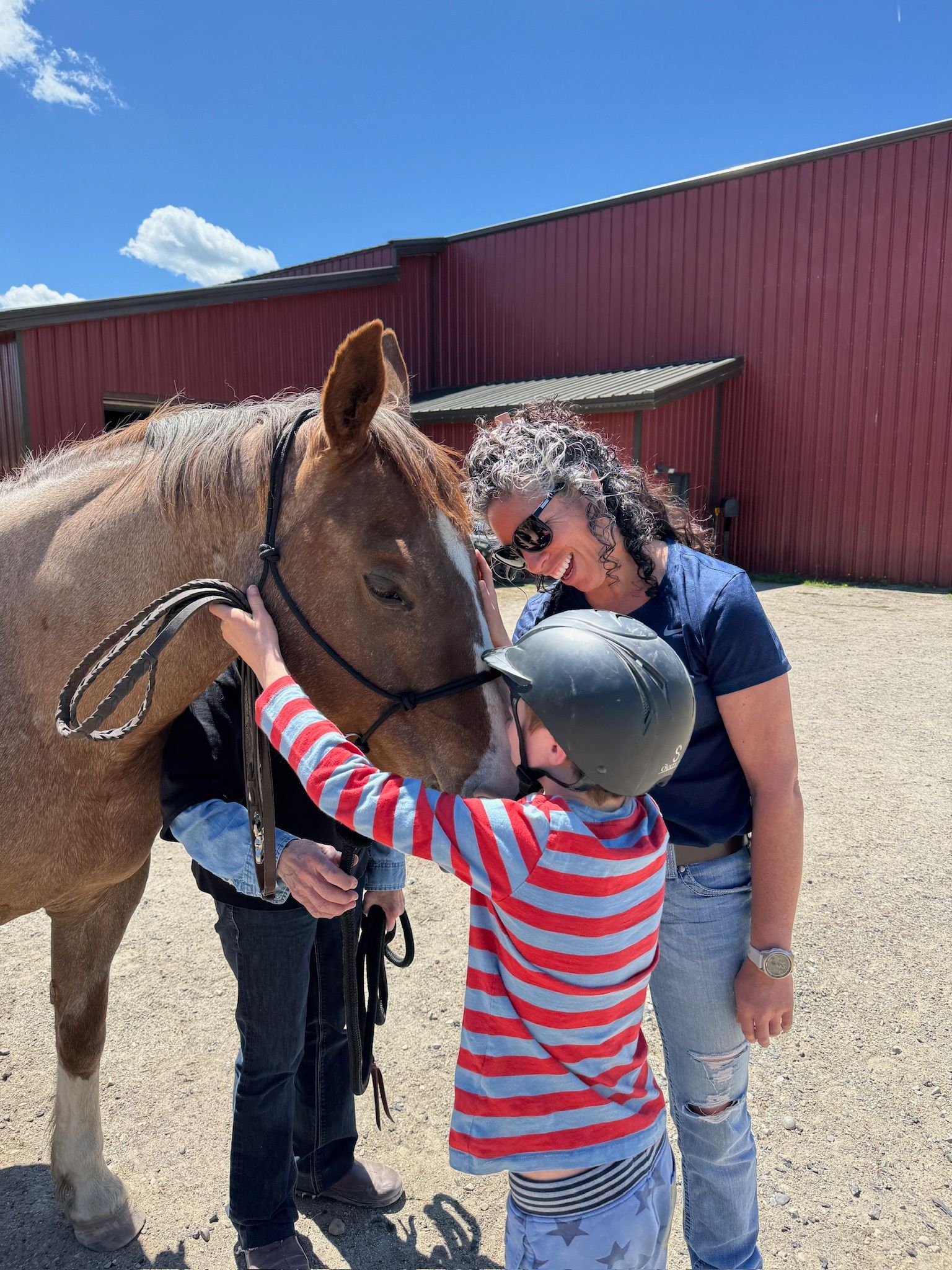 Child in helmet pets a horse while woman watches, outside a barn.
