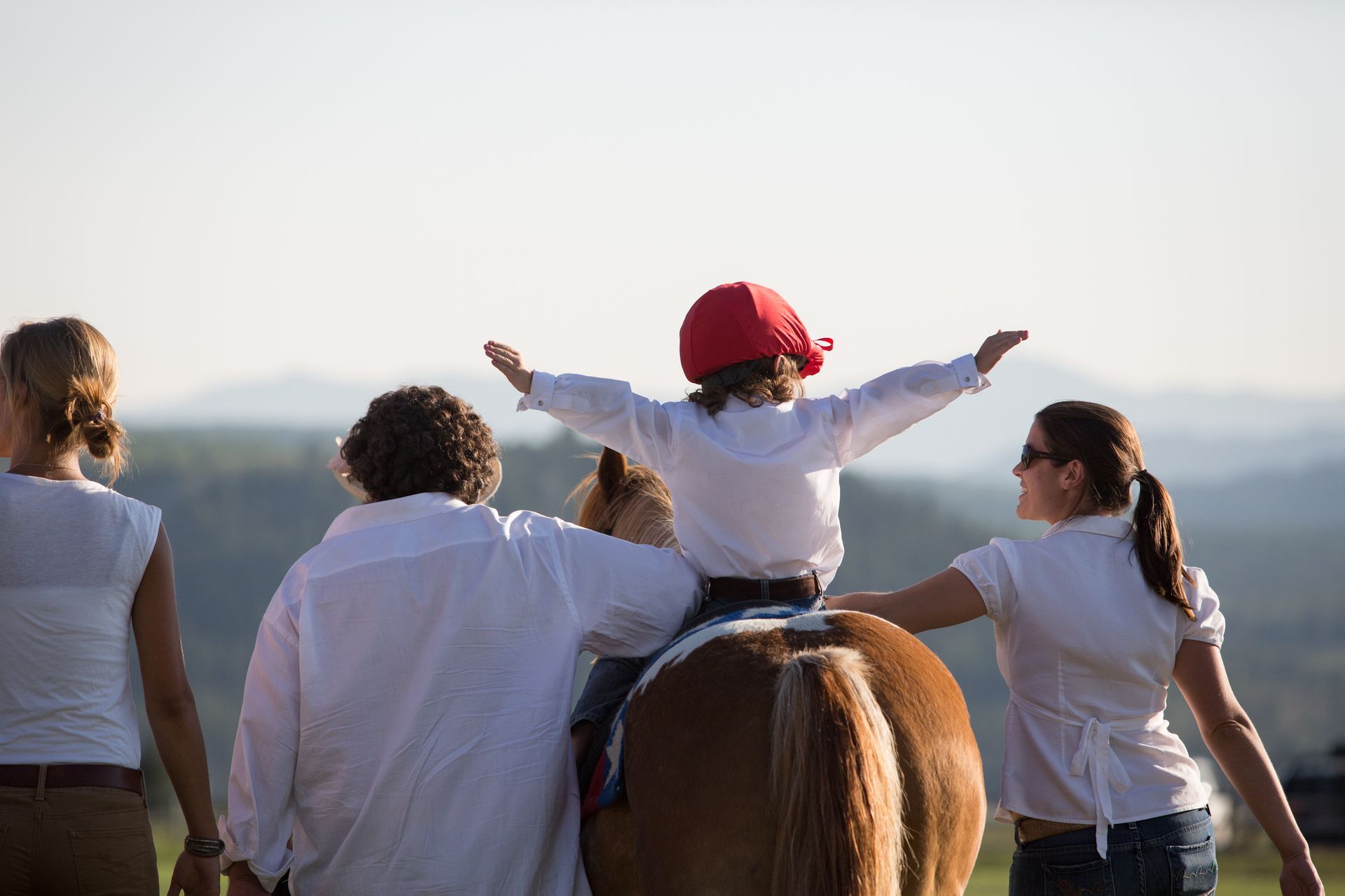 A person wearing a red cap sits on a horse with arms outstretched, flanked by two people in white shirts outdoors.