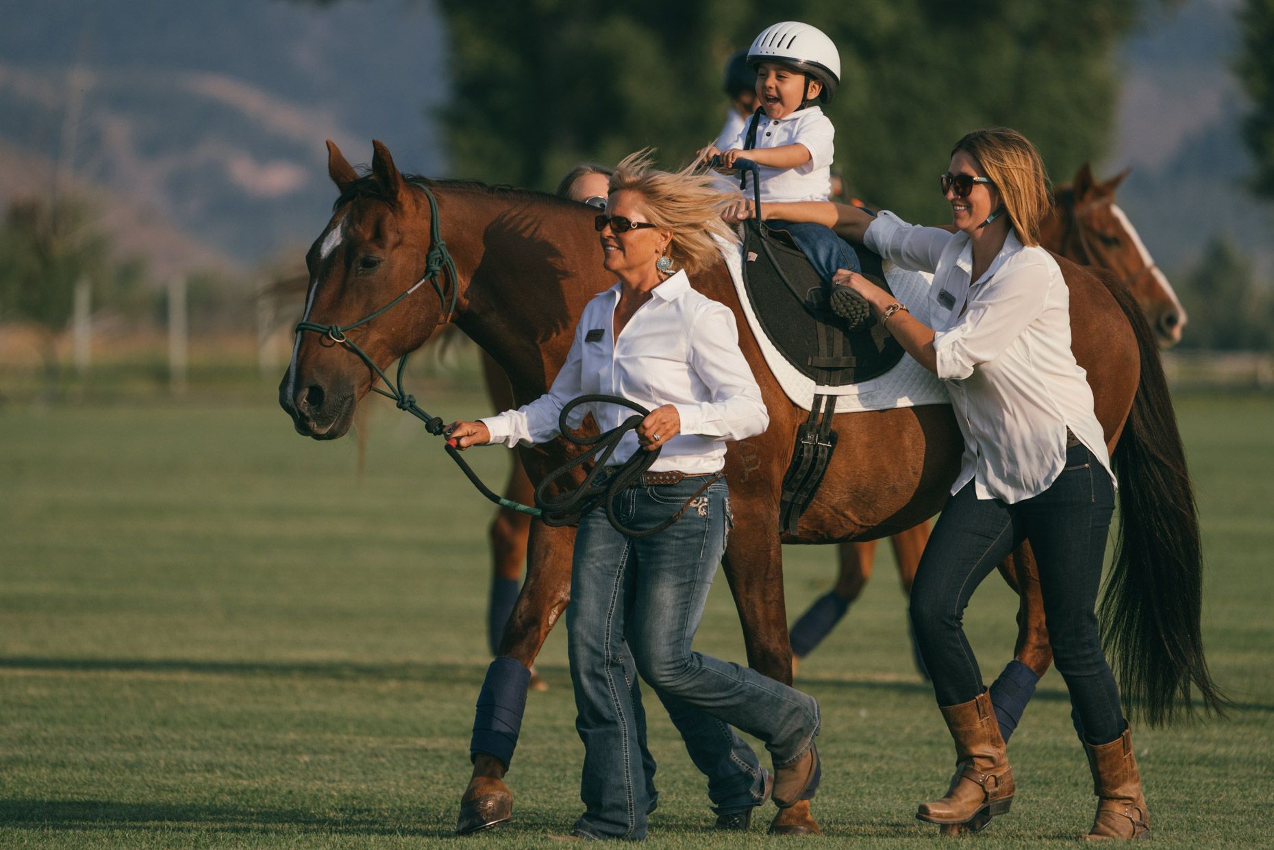 Two people in white shirts and jeans walk alongside a horse carrying a child in a helmet on a grassy field.