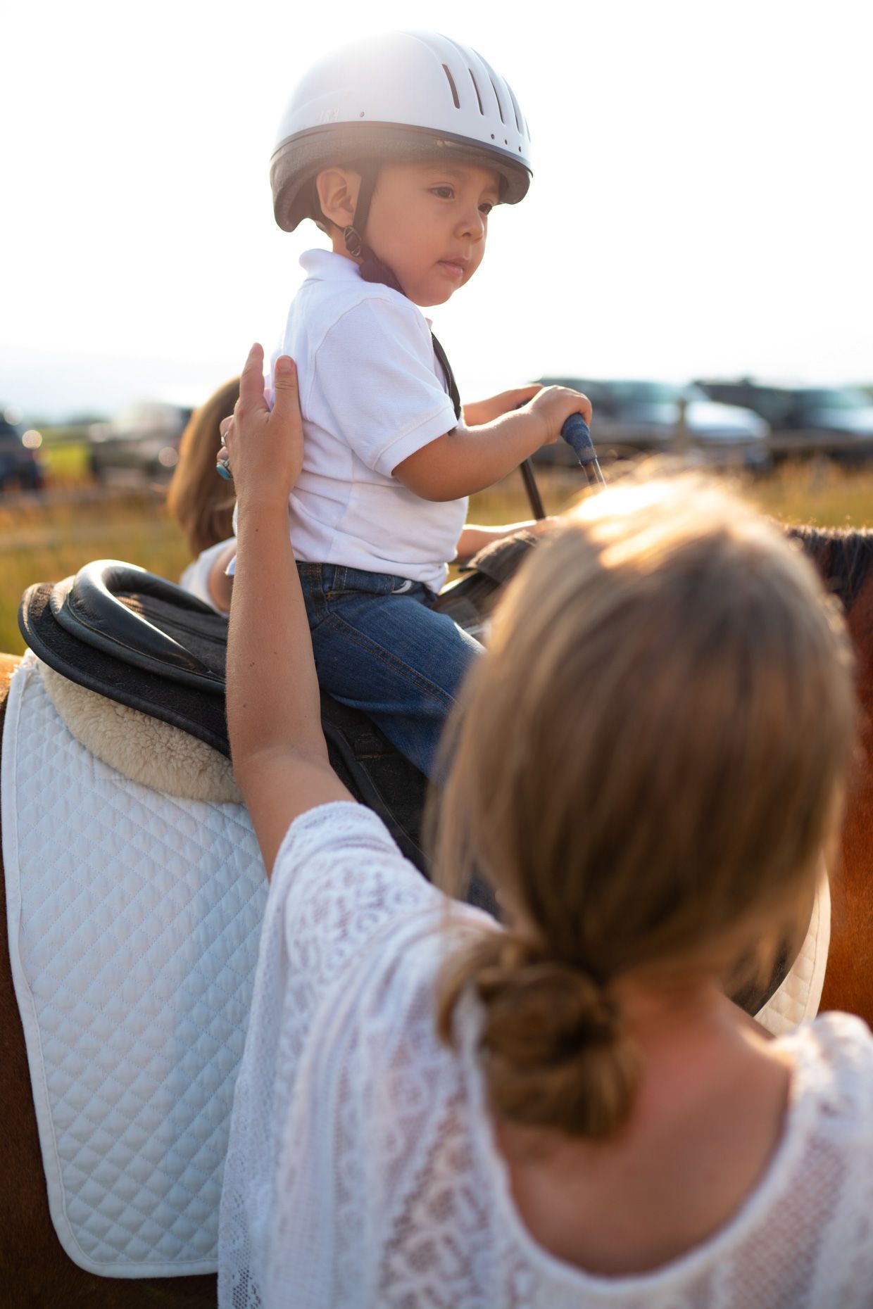 Child in helmet pets a horse while woman watches, outside a barn.