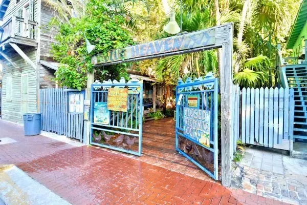 A blue gate stands at the entrance to Blue Heaven restaurant in Key West, framed by trees and a red brick sidewalk.