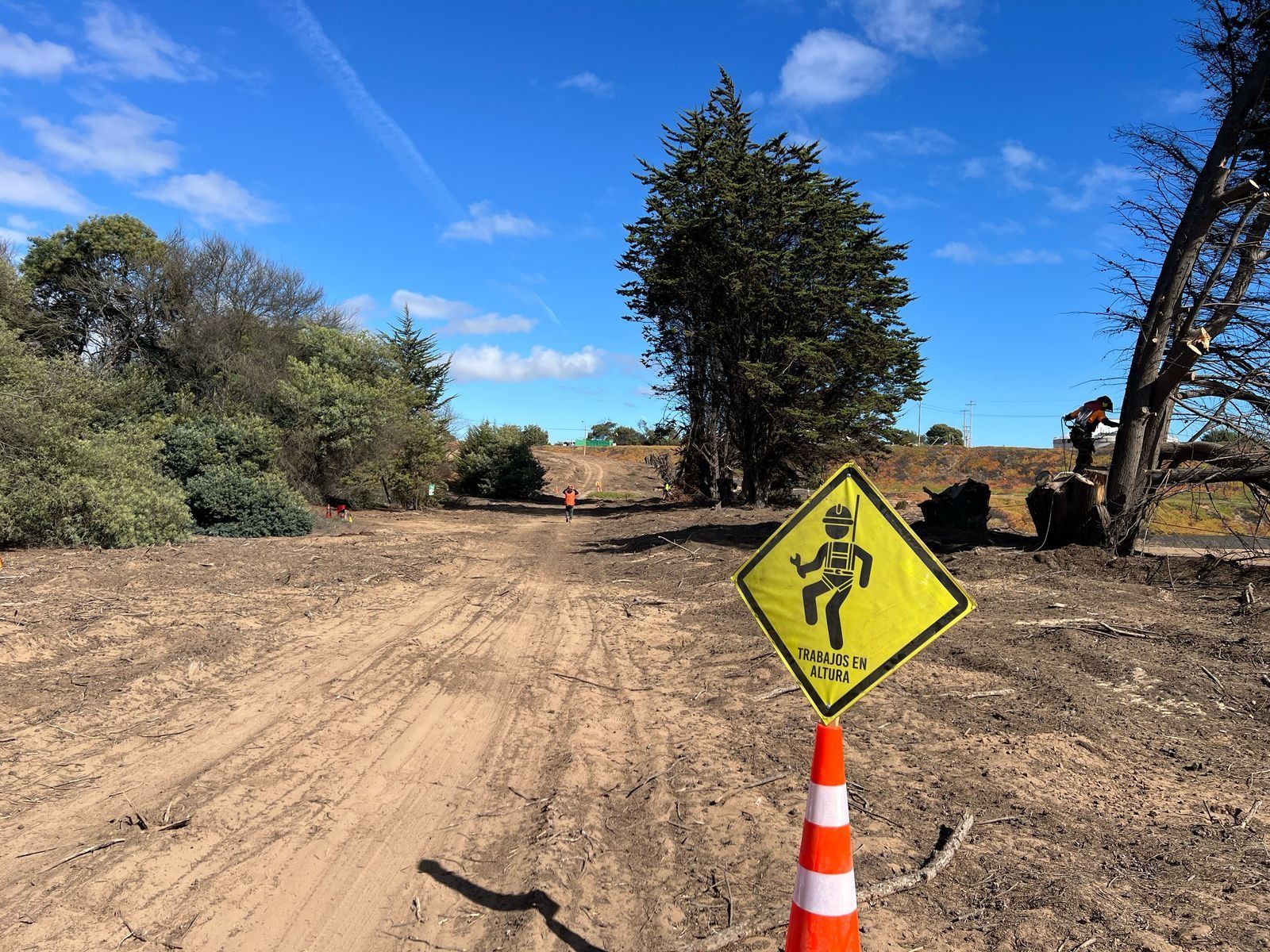 Un cartel amarillo con una persona está sobre un cono al costado de un camino de tierra.