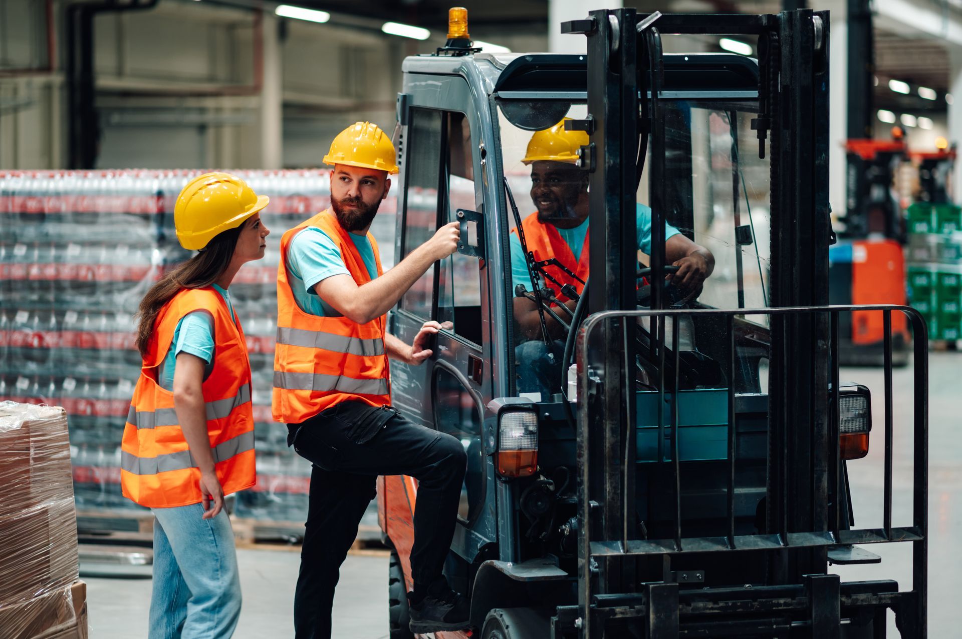 Three people in safety vests and hard hats in a warehouse; two standing, one operating a forklift.