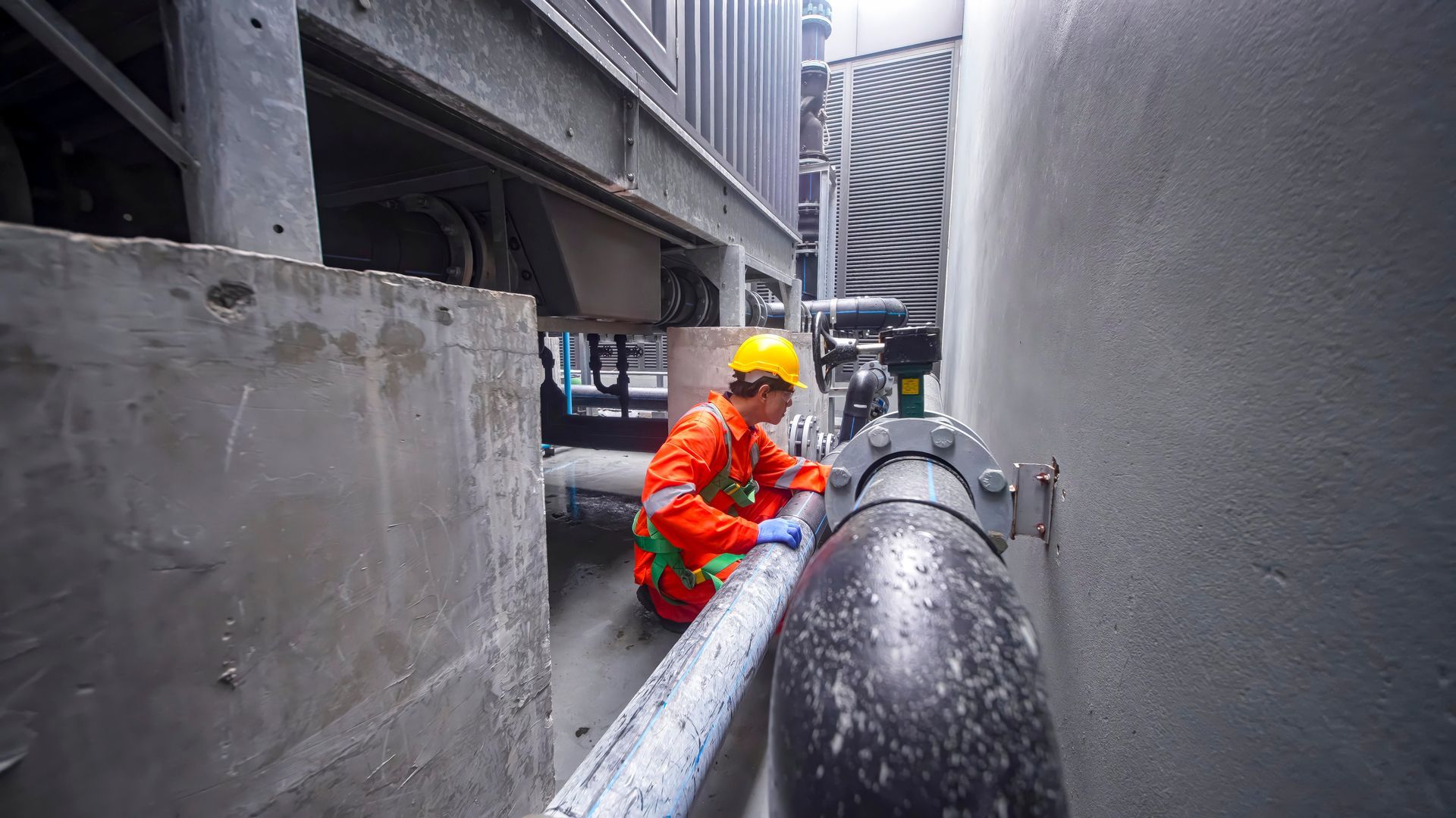 Worker in orange jumpsuit and hard hat inspects pipes between concrete structures.