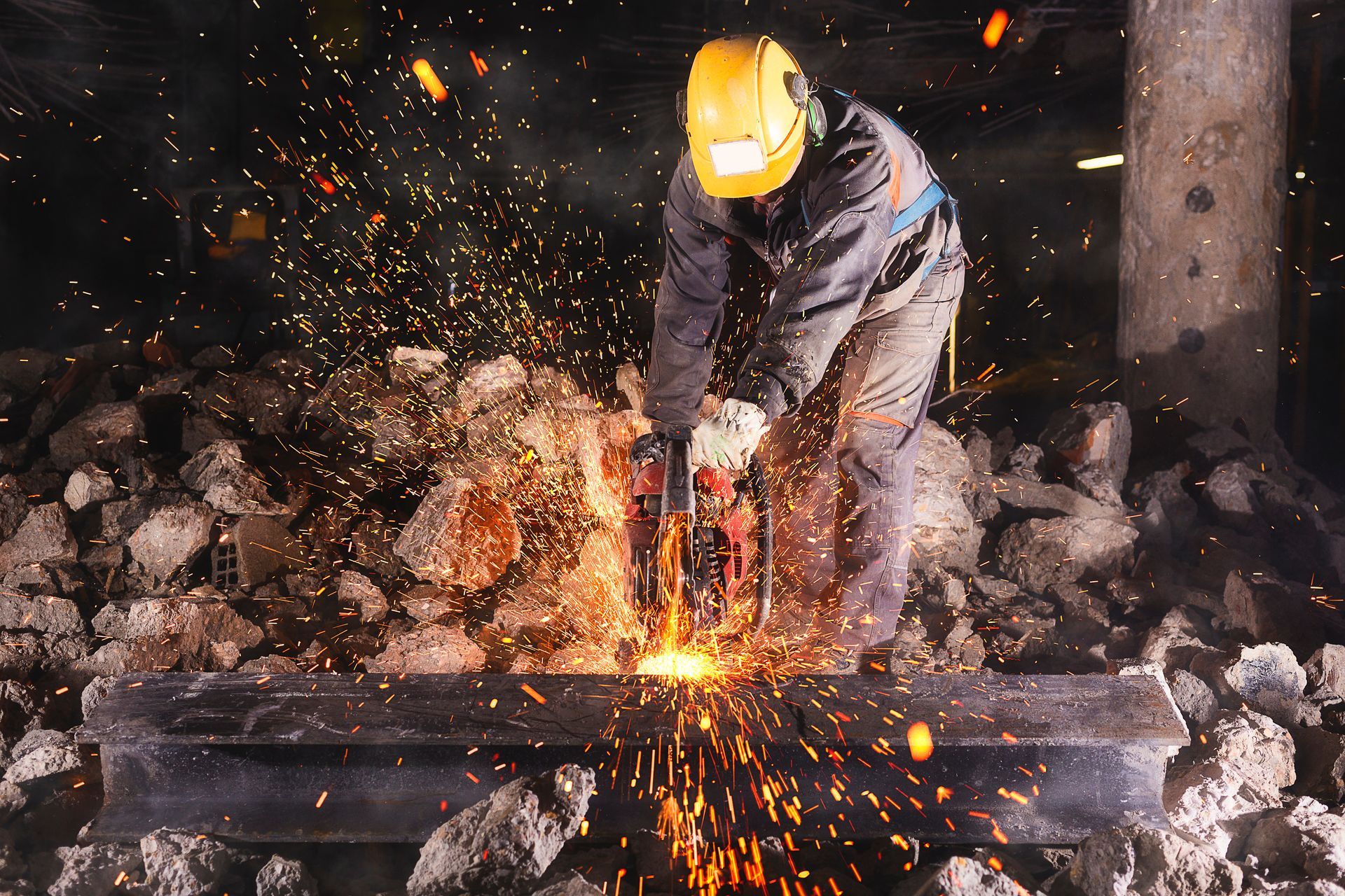 A worker in a helmet and protective gear uses a tool, creating sparks while cutting metal in a dark setting.