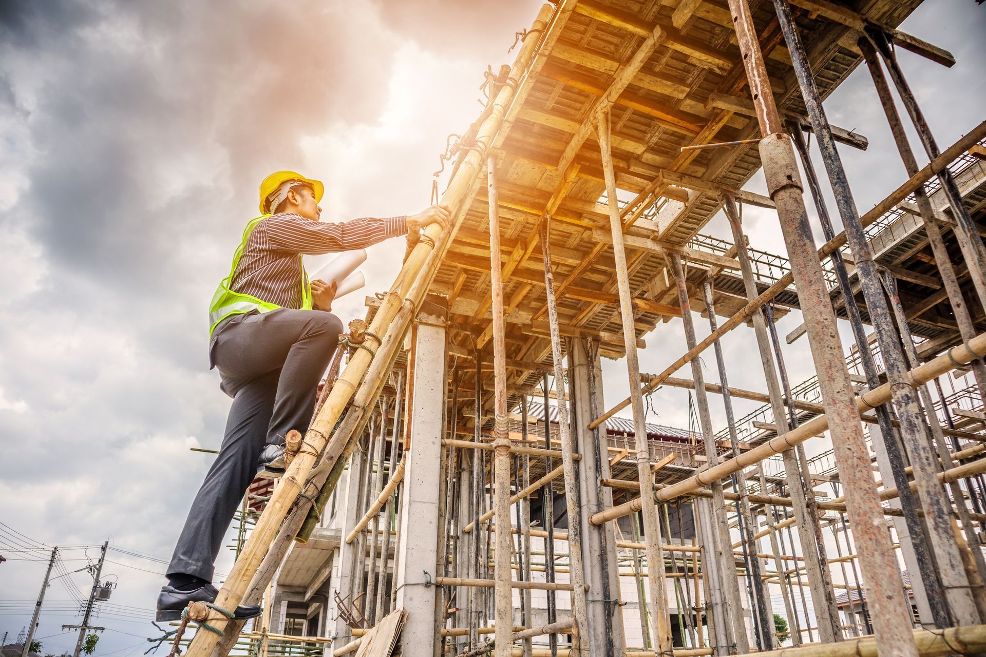 Construction worker climbs a wooden structure, wearing a hard hat and safety vest, under a cloudy sky.