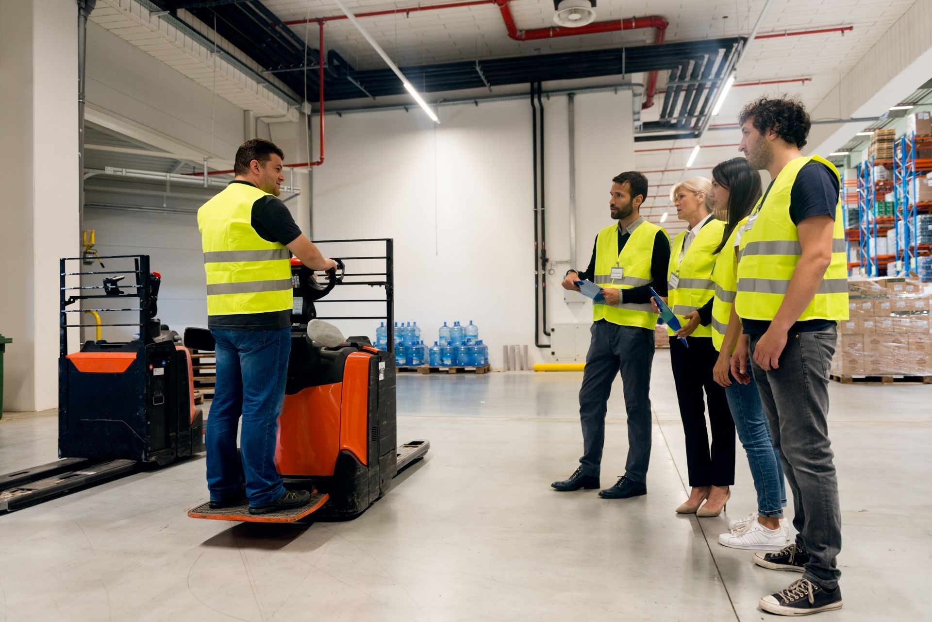 Warehouse worker demonstrates forklift operation to a group of people wearing safety vests.