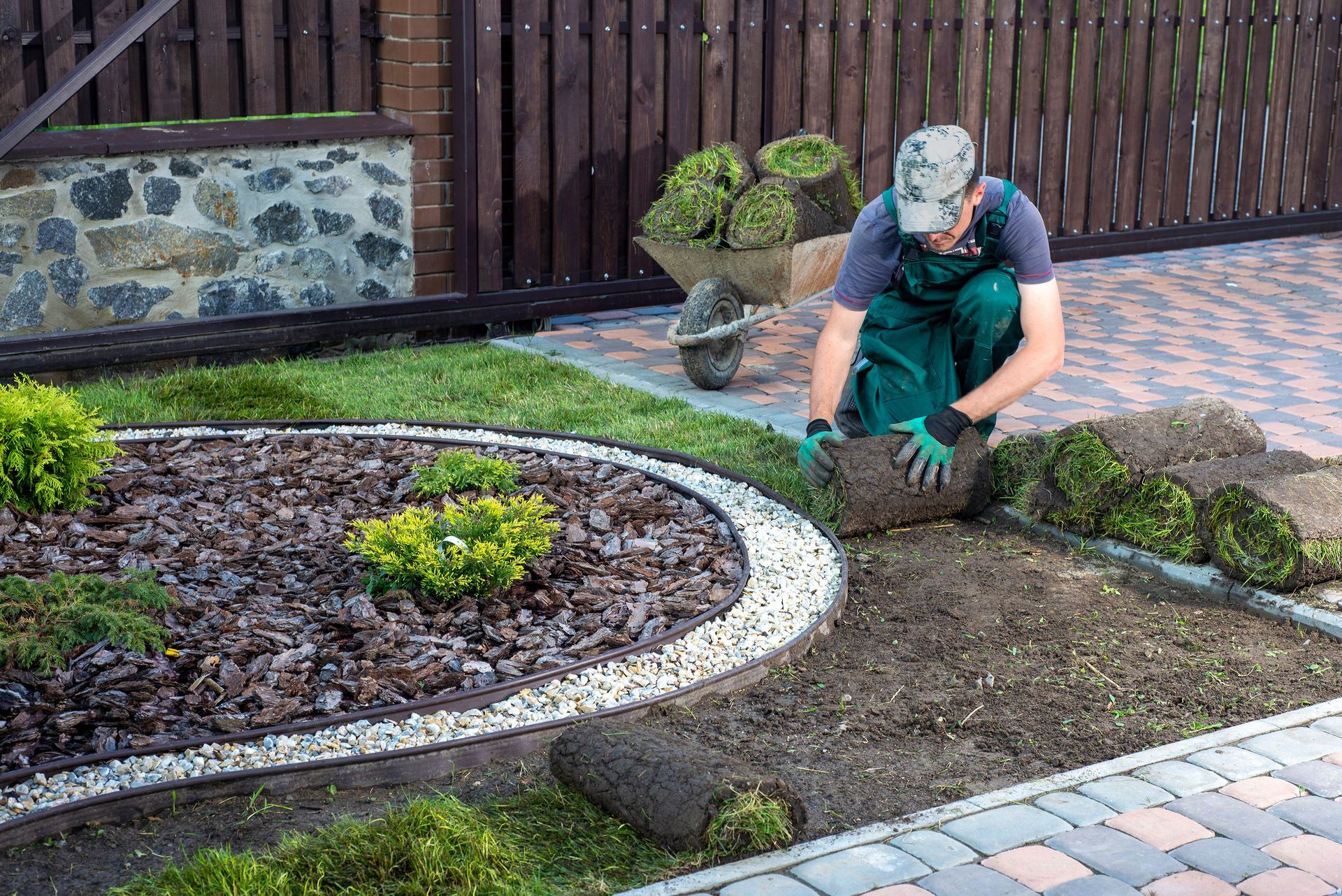 Man laying sod in a yard with a circular flower bed and a wheelbarrow nearby.