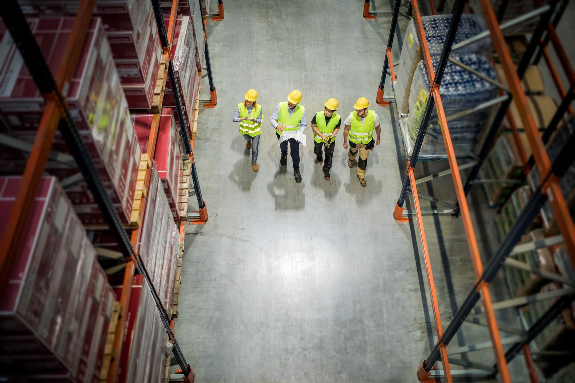 Warehouse workers in yellow vests and hard hats reviewing plans near shelves of goods.