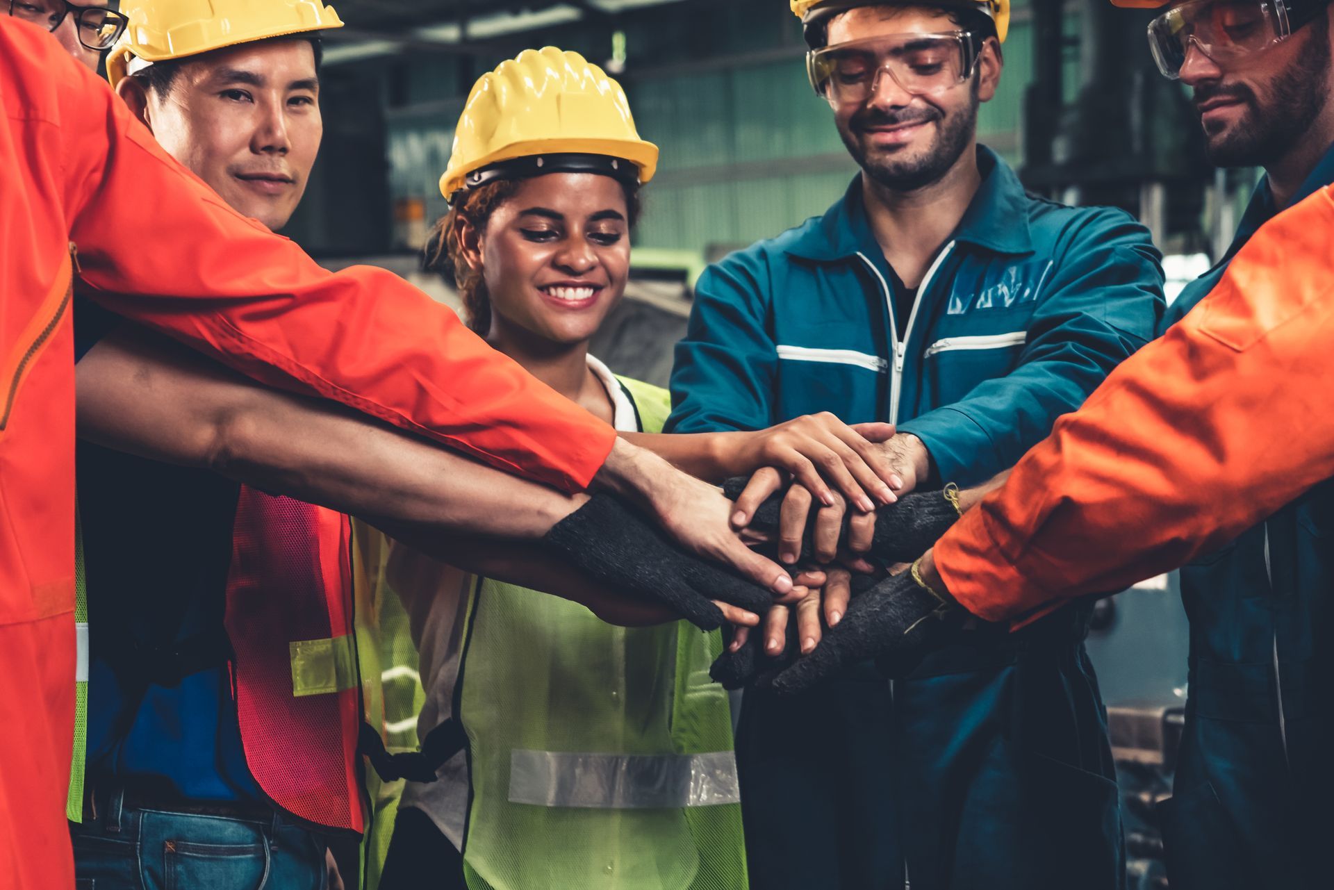 Factory workers in hard hats and safety gear with hands stacked together in a circle.