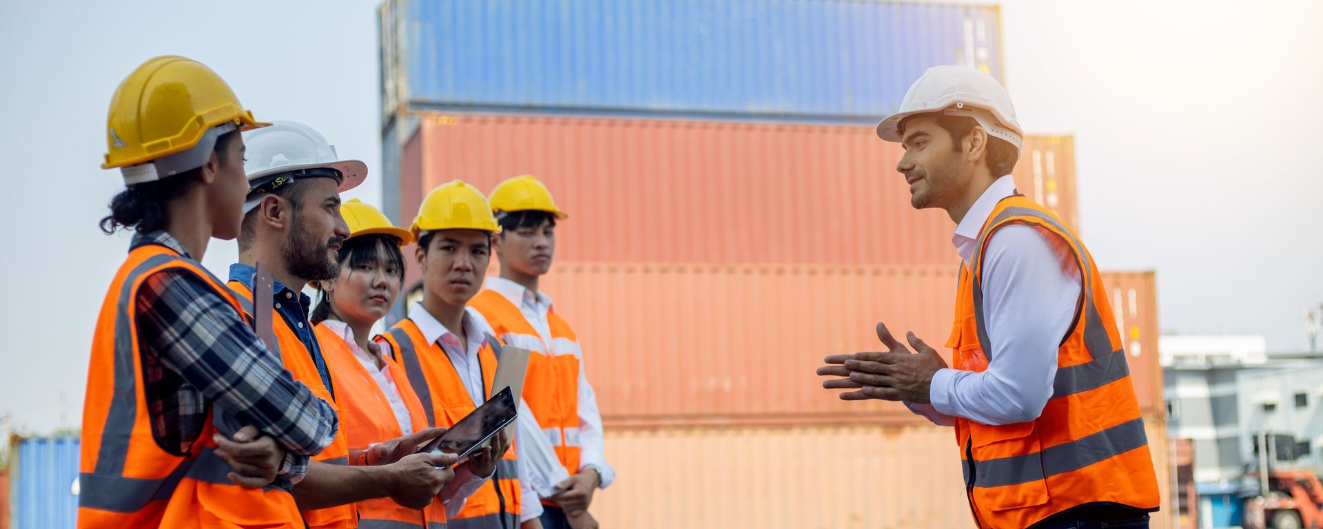 Group of workers in safety vests and hard hats at a shipping port, listening to a supervisor.