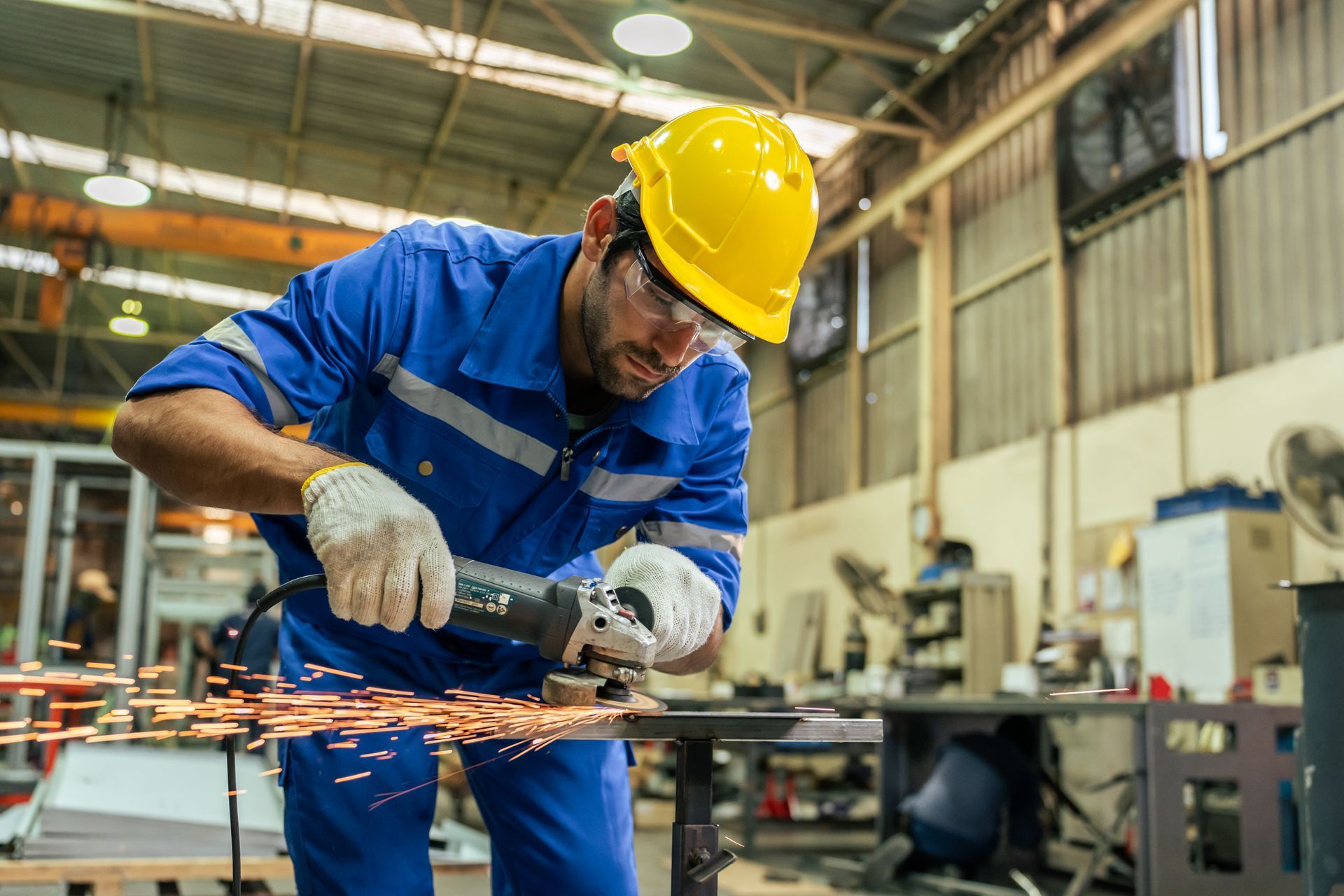 Person in blue work suit and hard hat using an angle grinder, sparks flying, inside a workshop. Person in blue work suit and hard hat using an angle grinder, sparks flying, inside a workshop.
