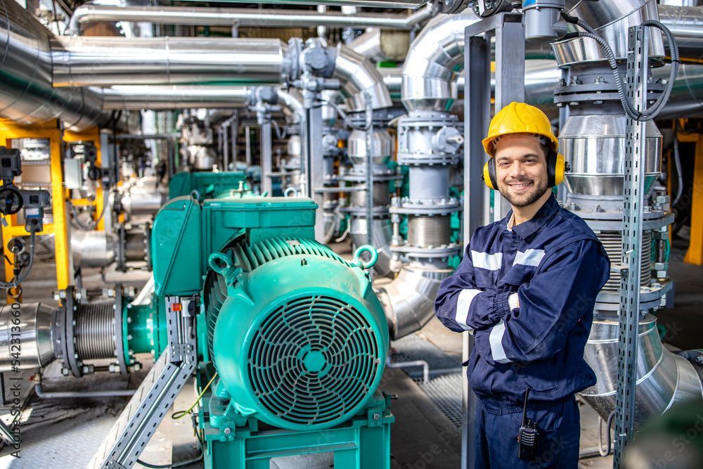 Engineer in a yellow hard hat and blue jumpsuit stands in a factory, arms crossed, smiling.