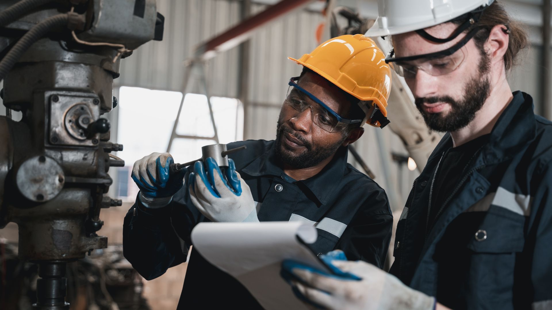 Two workers in safety gear examine a metal part, one with a clipboard, in a factory.