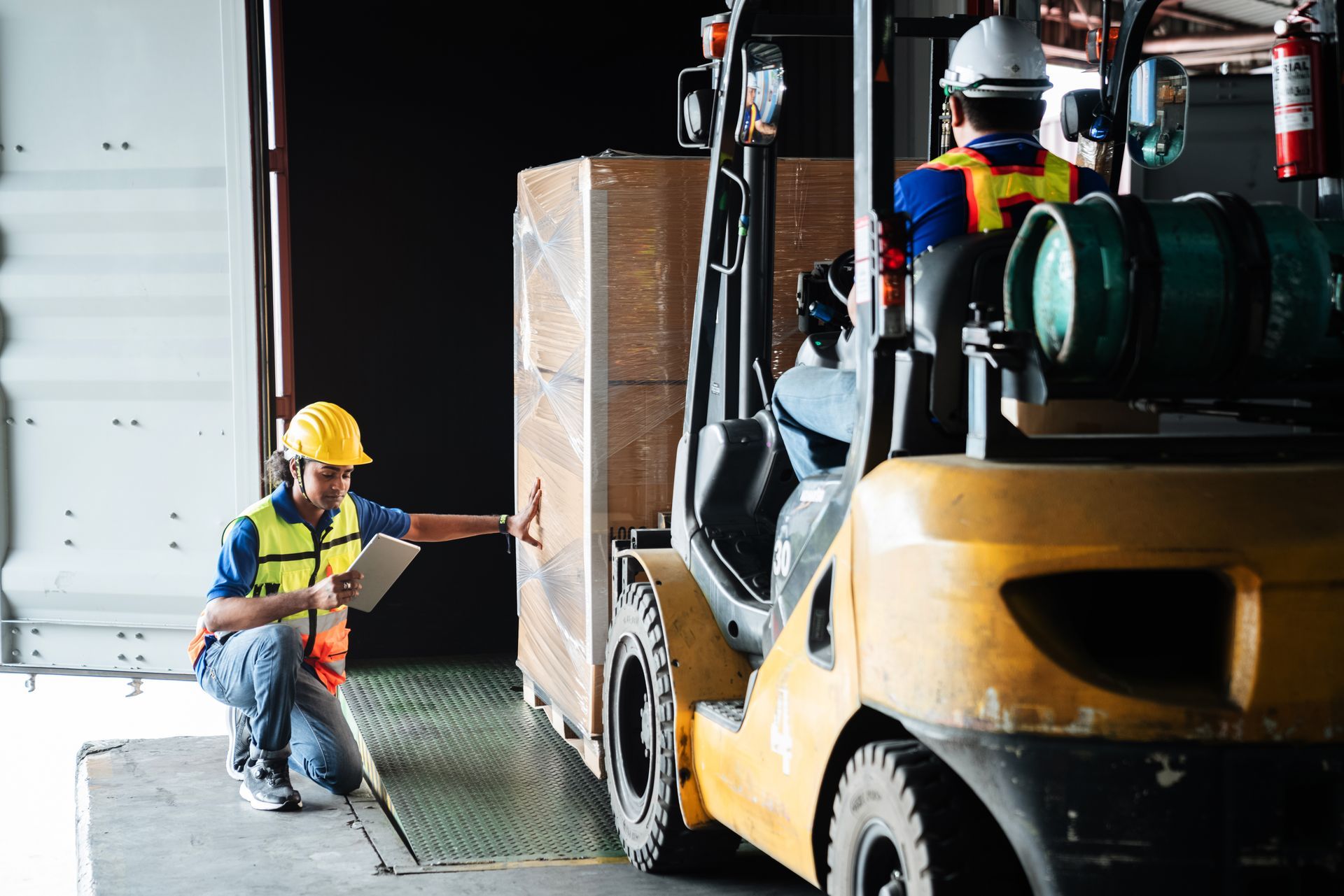 Warehouse workers loading crates with a forklift; one worker in a hard hat inspects.