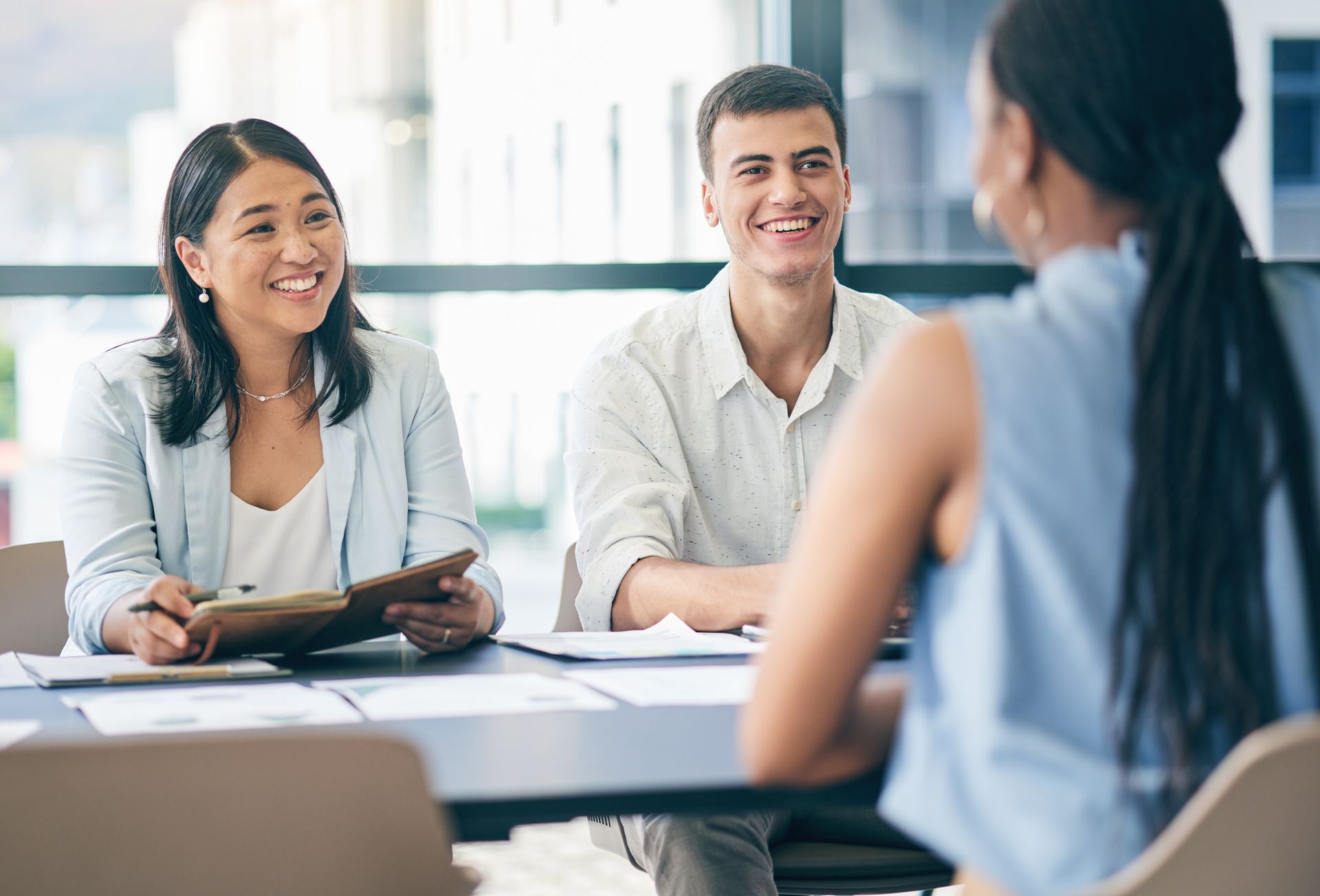 Three people smiling in a meeting; two interviewers, one interviewee, at a table with papers.