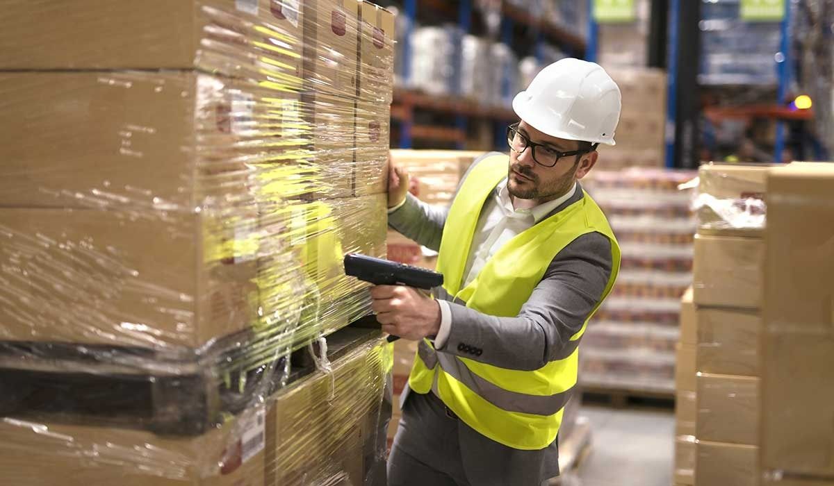 Warehouse worker scans boxes on a pallet, wearing a hard hat and safety vest.