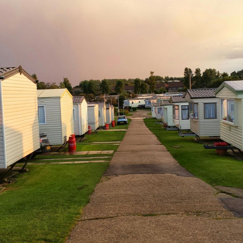 Row of white trailer homes with a central path, green grass, and a soft sunset sky.