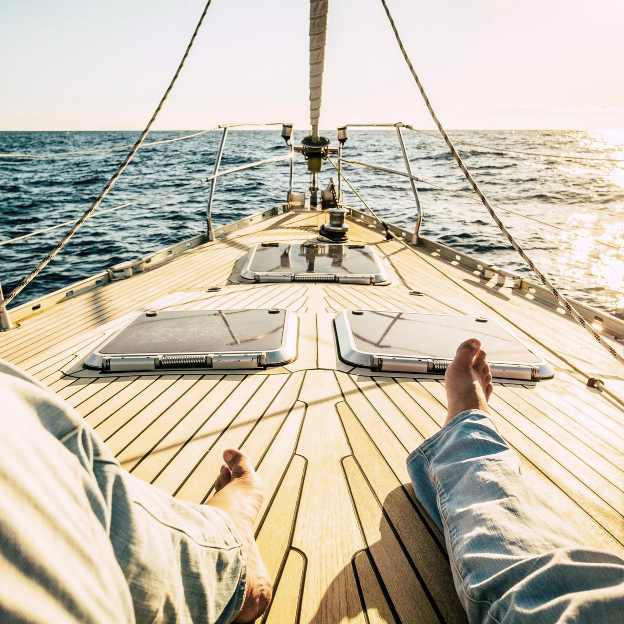 Person's bare feet and legs on a wooden sailboat deck, sunlit ocean view.